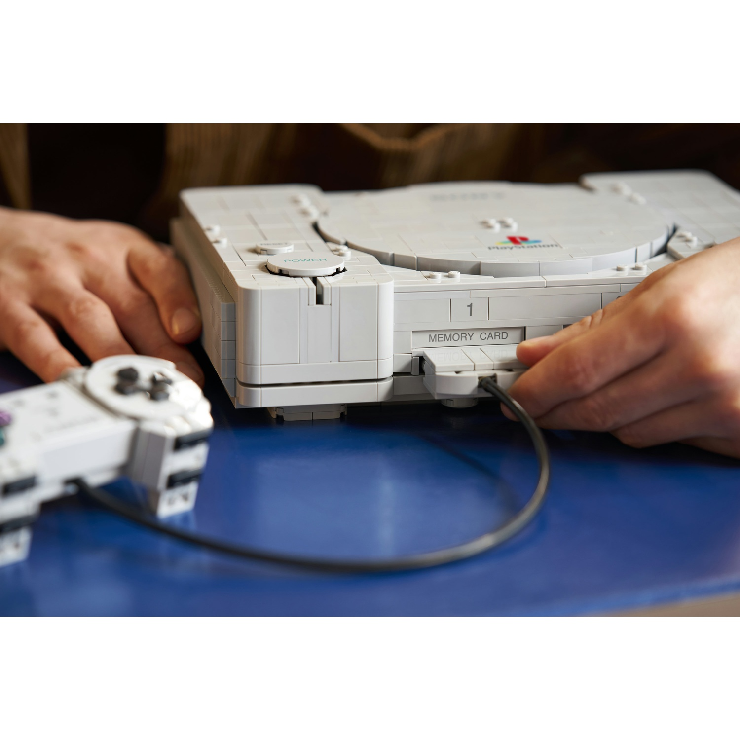 Una persona collega un controller a una console di gioco Lego grigia su un tavolo blu.