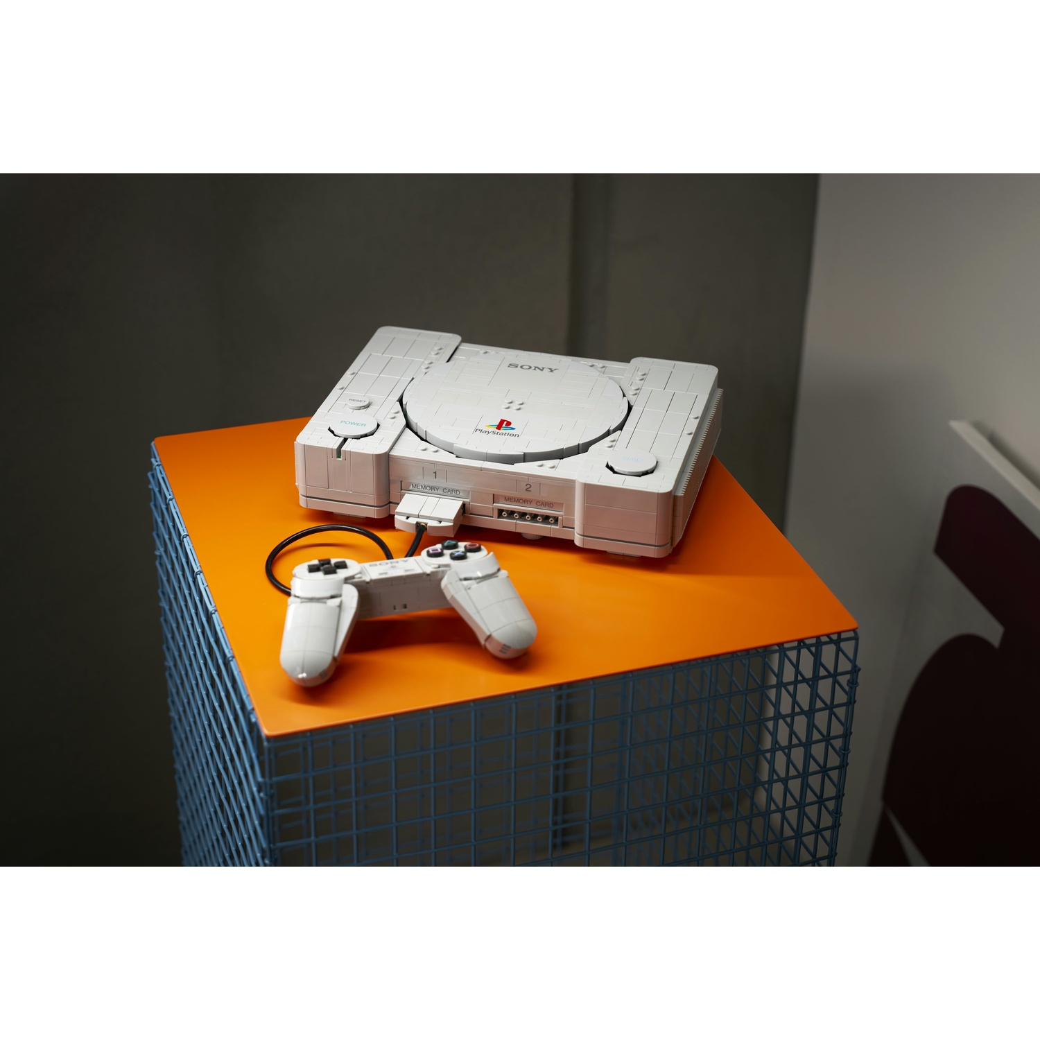 Una console PlayStation 1 grigia in mattoncini con controller coordinato si trova su una superficie arancione.