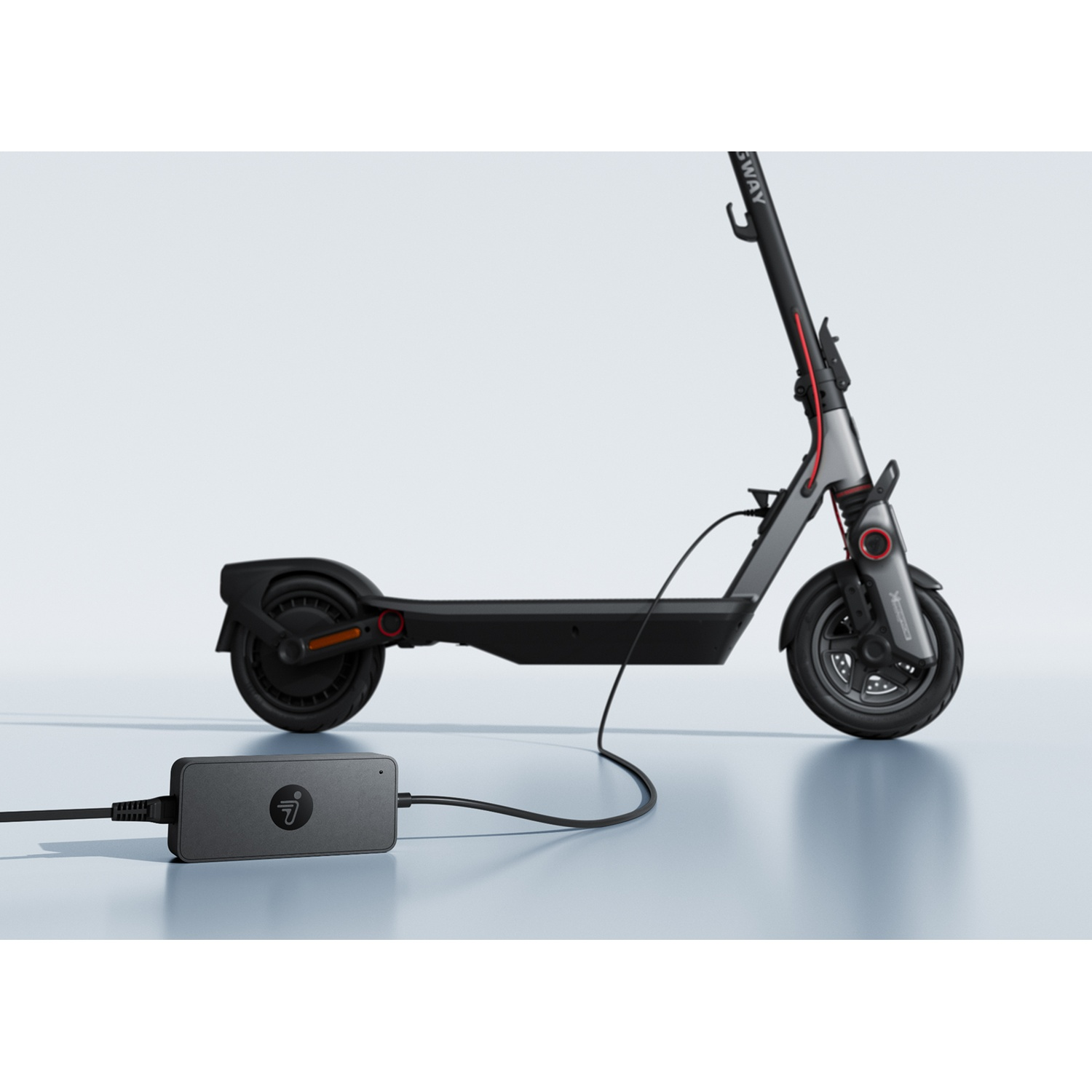 Une trottinette électrique grise se tient sur une surface claire et est reliée à un chargeur noir.