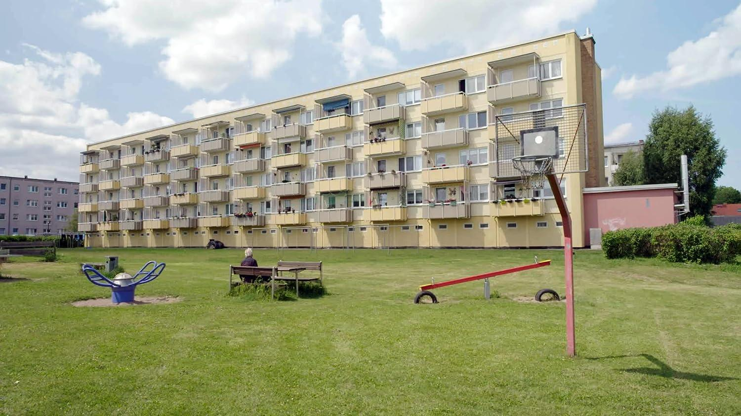 Eine grüne Wiese mit Spielplatz und Basketballkorb vor einem großen gelben Wohnblock mit vielen Balkonen.
