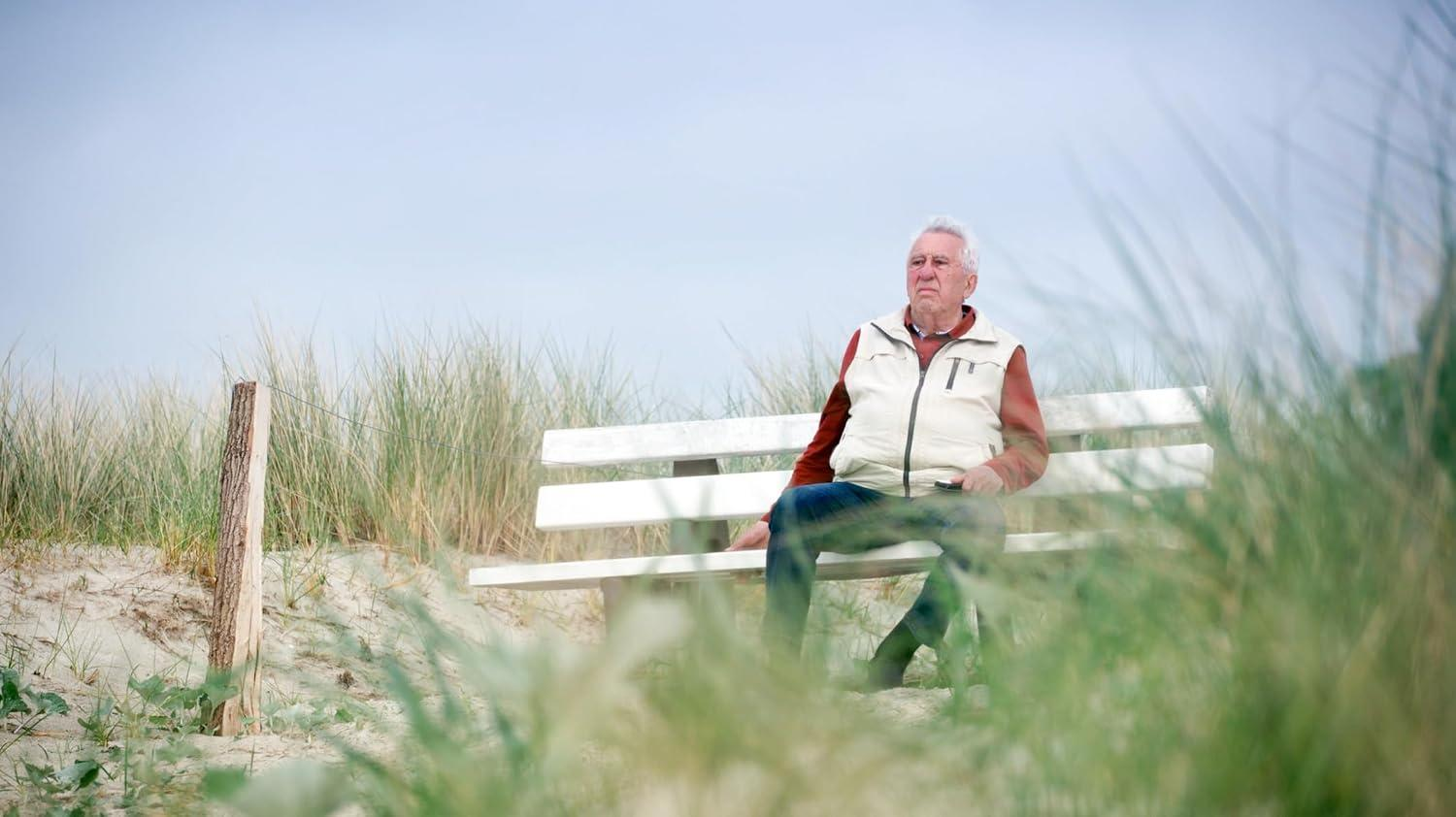 Älterer Mann sitzt auf einer weißen Holzbank im Gras in der Nähe von Sanddünen unter bewölktem Himmel.
