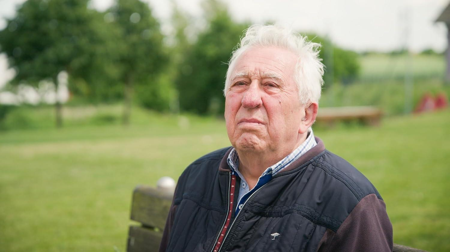 Älterer Mann mit weißem Haar sitzt in einem Park und blickt nachdenklich in die Ferne.