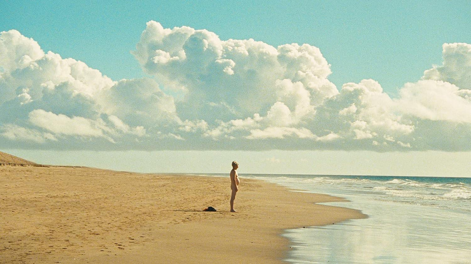 Eine Person steht allein an einem sandigen Strand und blickt unter dramatischem Himmel auf das Meer.