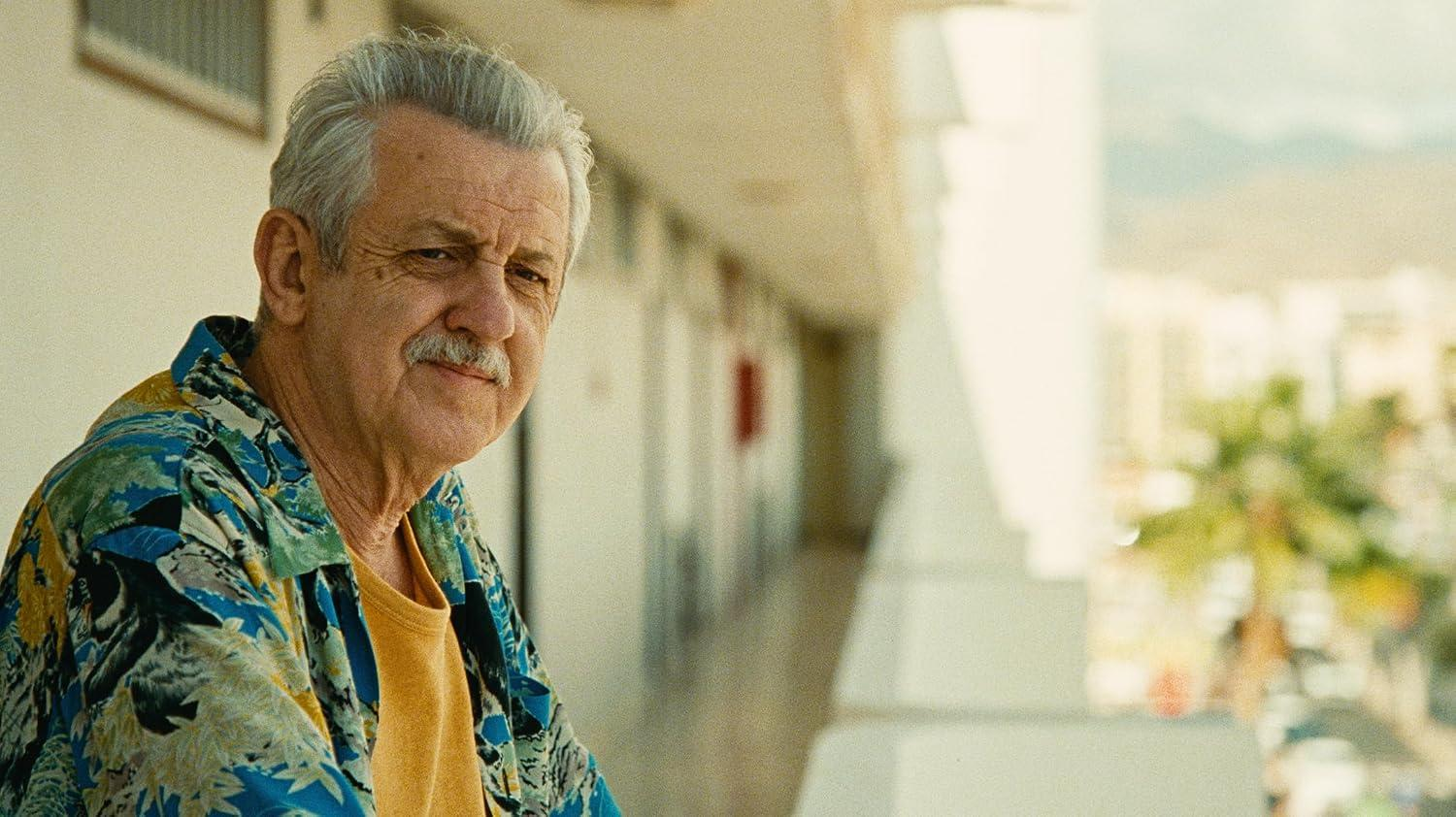 Älterer Mann mit grauem Haar und Schnurrbart in einem bunten Hemd, auf einem sonnigen Balkon.