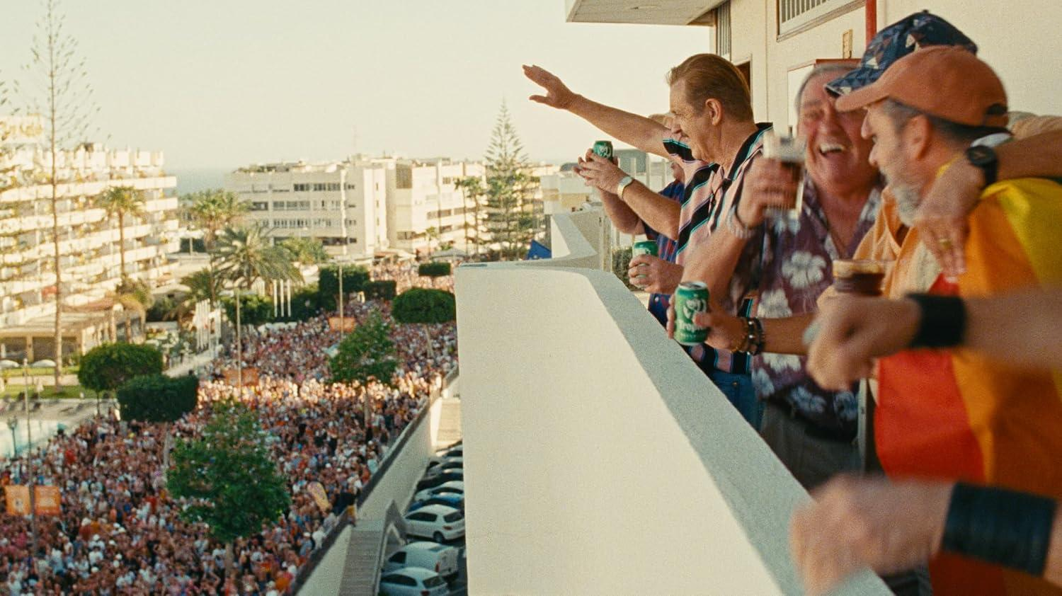 Männer auf einem Balkon erheben Getränke, winken und lachen an einem sonnigen Tag in eine Menge.