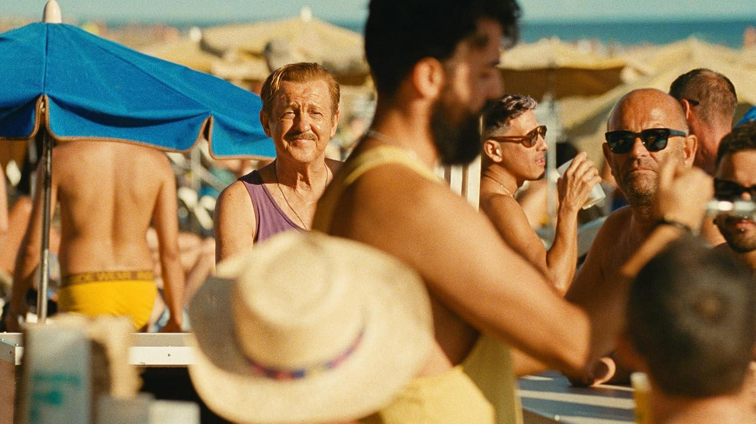 Menschen entspannen an einem sonnigen Strand, mit einem blauen Sonnenschirm links und Strandbesuchern im Hintergrund.