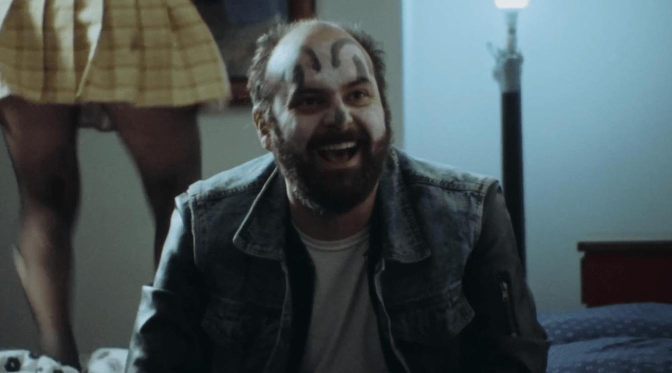 Ein glücklicher Mann mit Gesichtsbemalung sitzt in einem Raum, während im Hintergrund eine Person in einem gelben Rock steht.