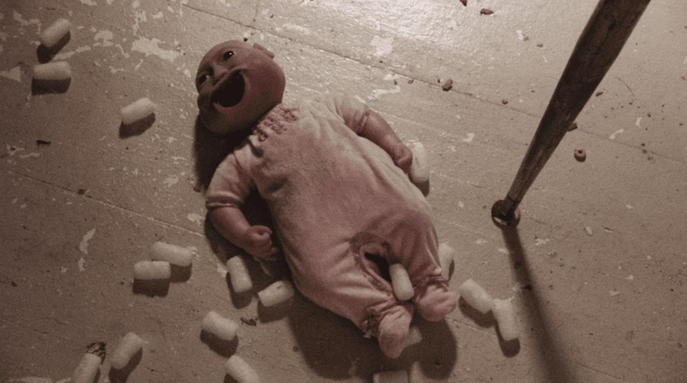 Eine gruselige Puppe in pinker Kleidung liegt auf einem Holzboden, umgeben von Verpackungsmaterial.
