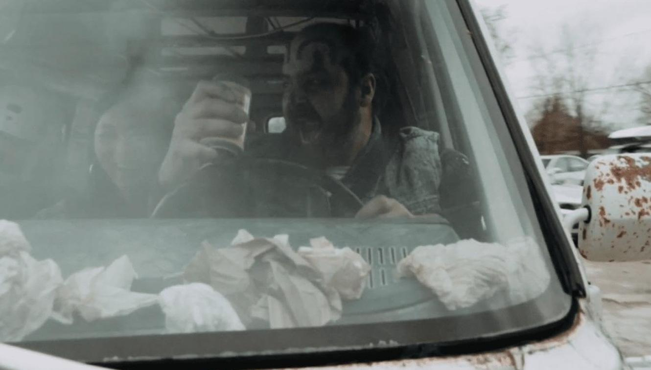 Ein Mann mit Gesichtsbemalung sitzt im Auto und hält ein Getränk, im Hintergrund ist eine Person zu sehen.