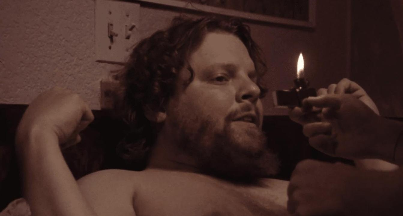 Ein bärtiger Mann liegt im Bett, während jemand eine brennende Kerze vor ihn hält.