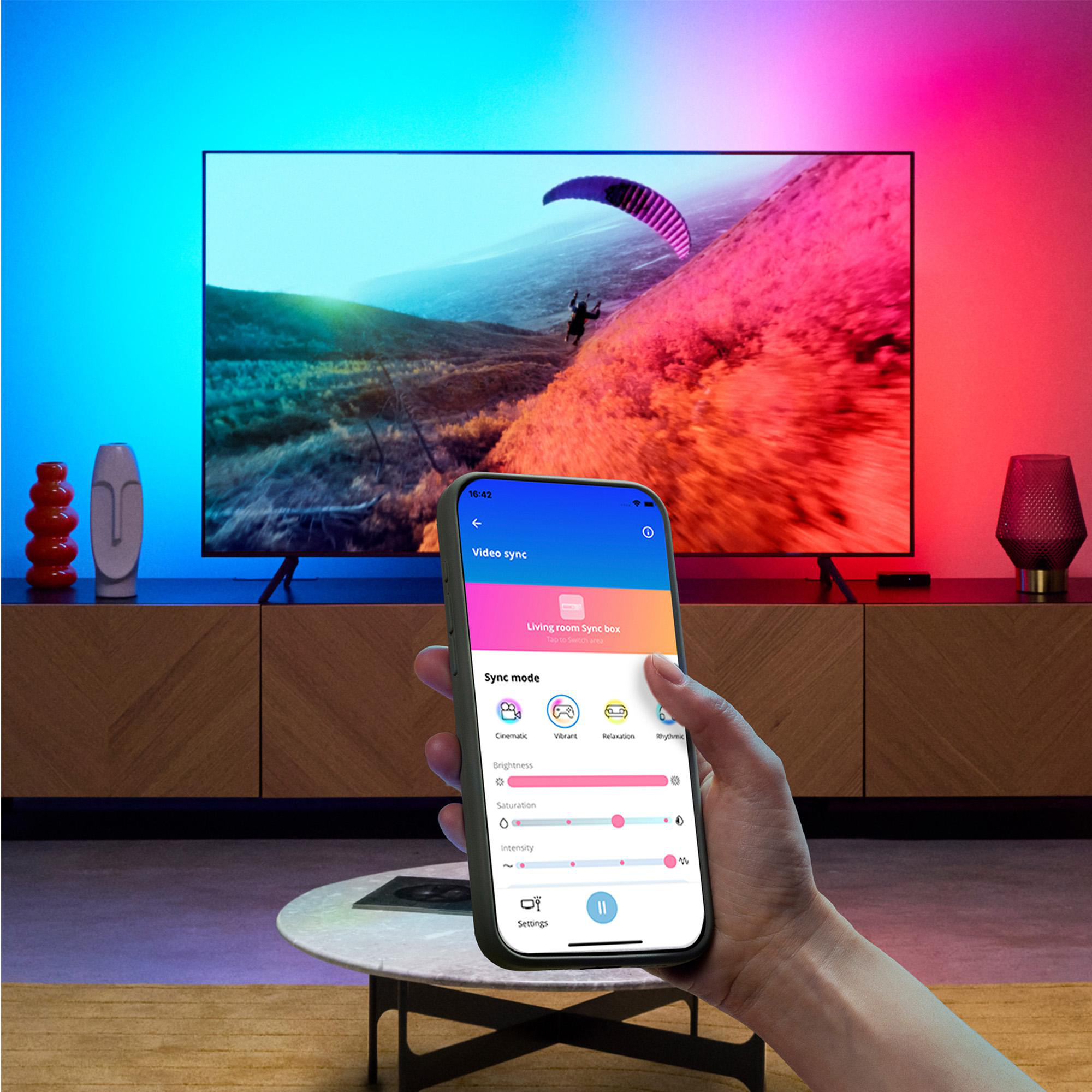 Hand hält Smartphone mit Video-Sync-App, im Hintergrund ein Fernseher mit farbigen Lichtern.