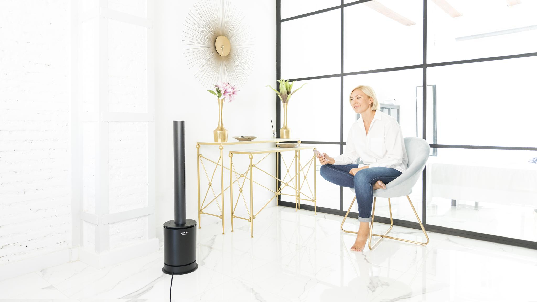 Mujer rubia se sienta en una habitación moderna de suelo blanco en un sillón azul claro junto a un ventilador de torre negro.