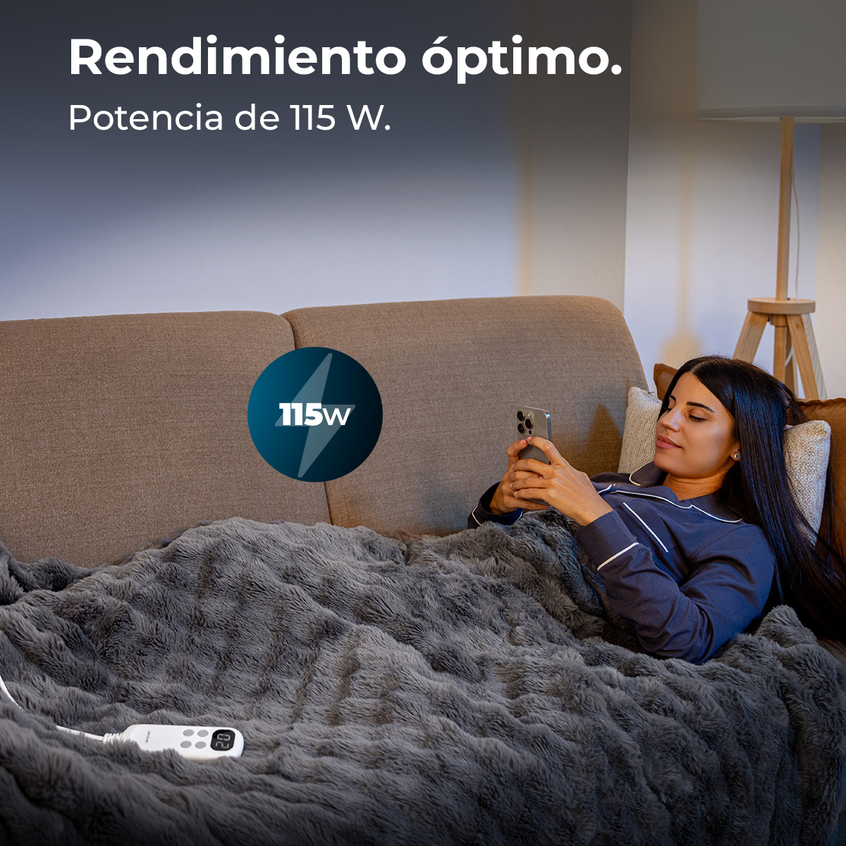 Una mujer se relaja en un sofá bajo una manta gris peluda, usando su teléfono inteligente.