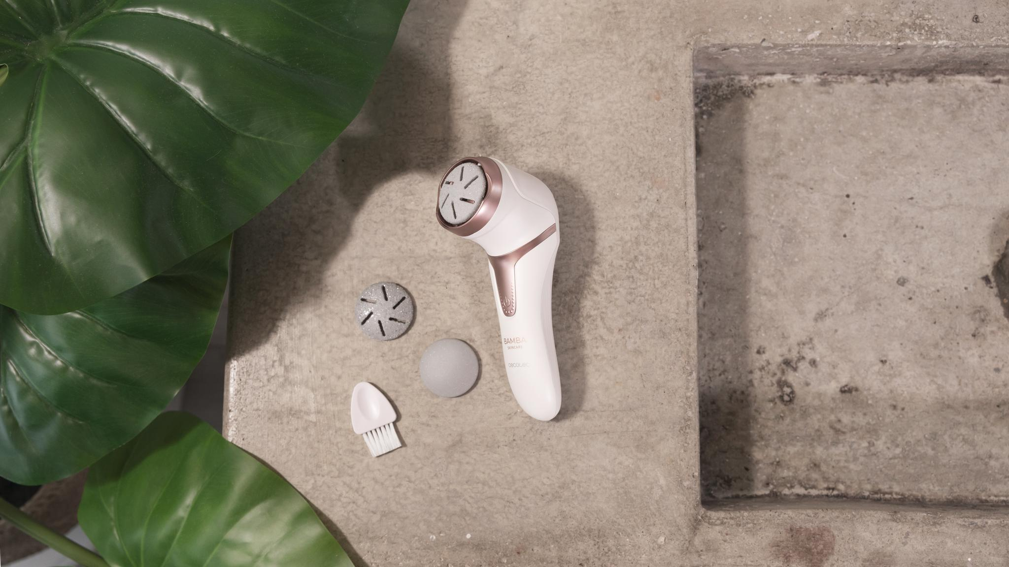 Un dispositivo de limpieza facial blanco y oro rosa con accesorios yace sobre una superficie gris junto a hojas grandes.