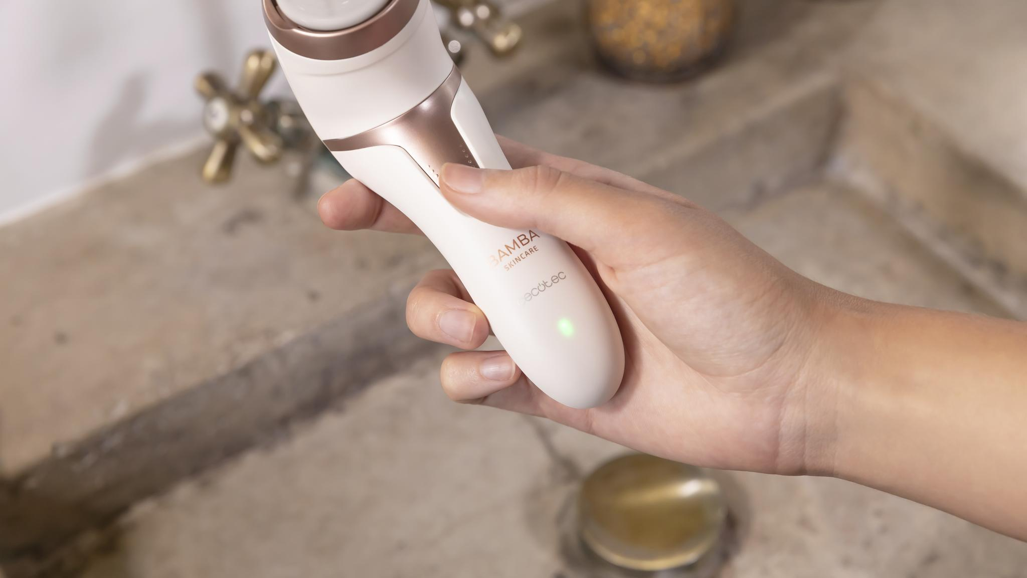 Una mano sostiene un dispositivo de cuidado facial blanco y dorado sobre un lavabo.