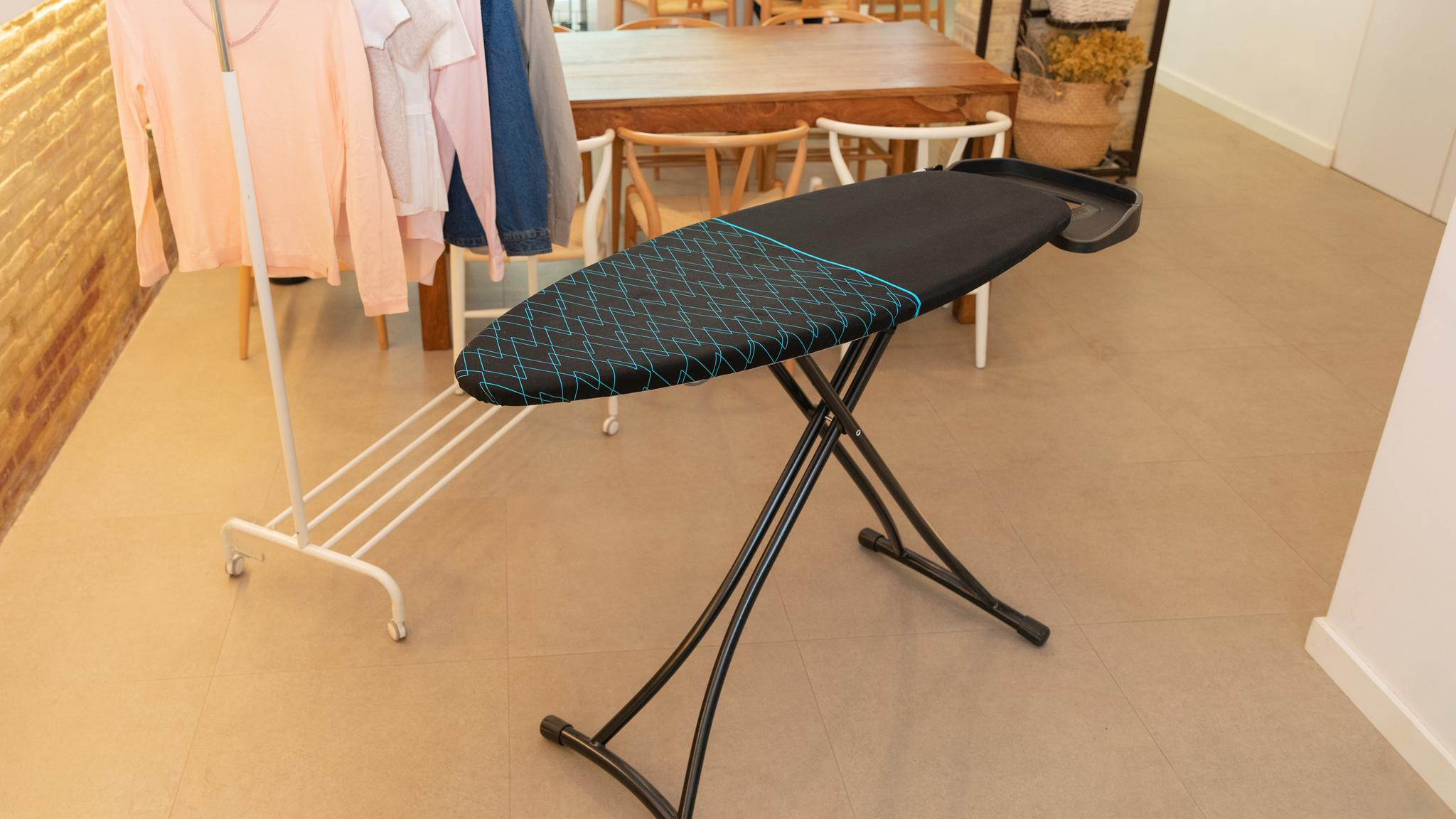 Una tabla de planchar negra con patrón en zigzag azul en una habitación, con perchero y mesa de comedor al fondo.