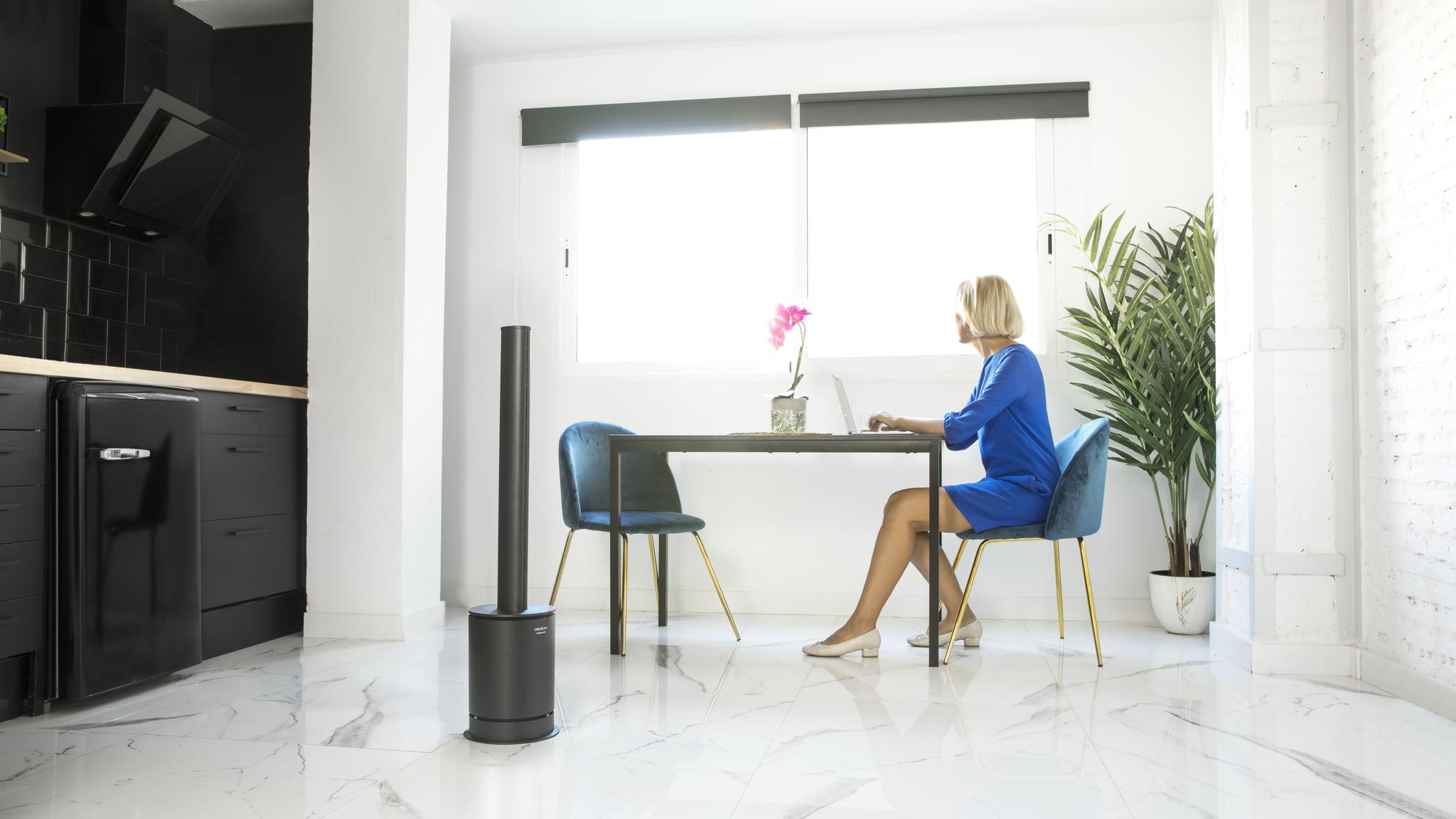 Una mujer con vestido azul se sienta a una mesa junto a una ventana en una habitación moderna con un ventilador.