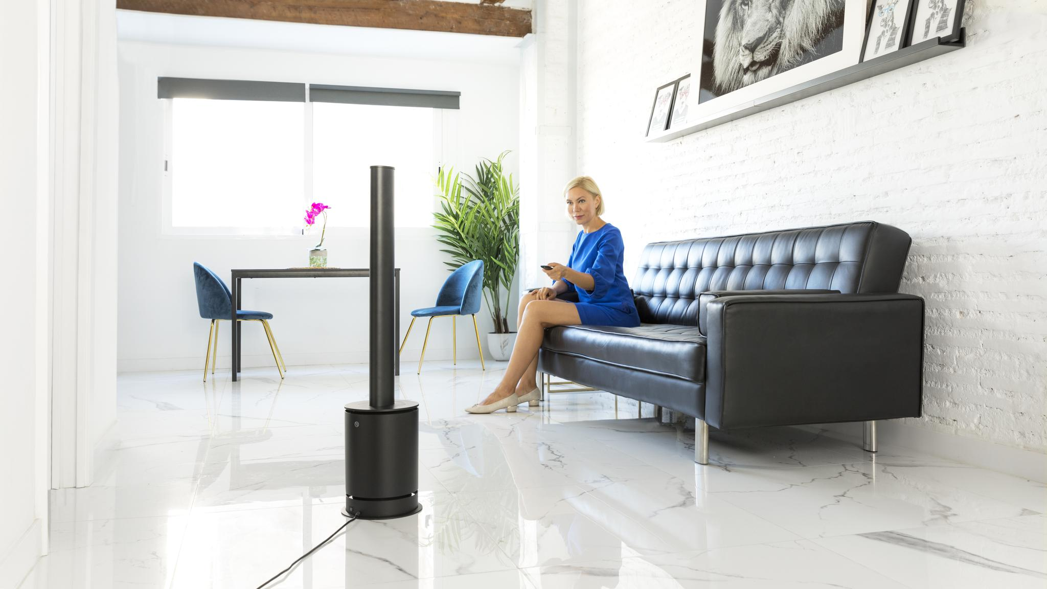 Una mujer rubia se sienta en un sofá negro en una habitación moderna, sosteniendo un control remoto.