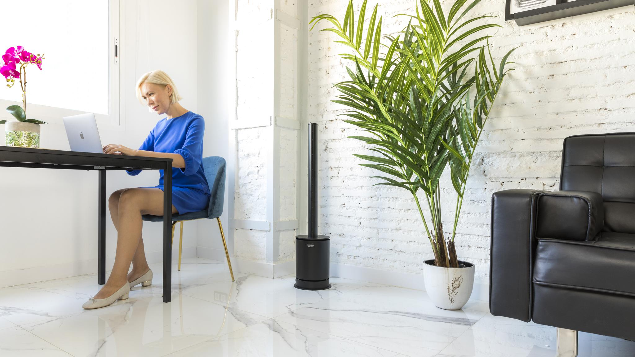 Una mujer rubia con vestido azul está sentada en una mesa trabajando en un portátil en una habitación luminosa.