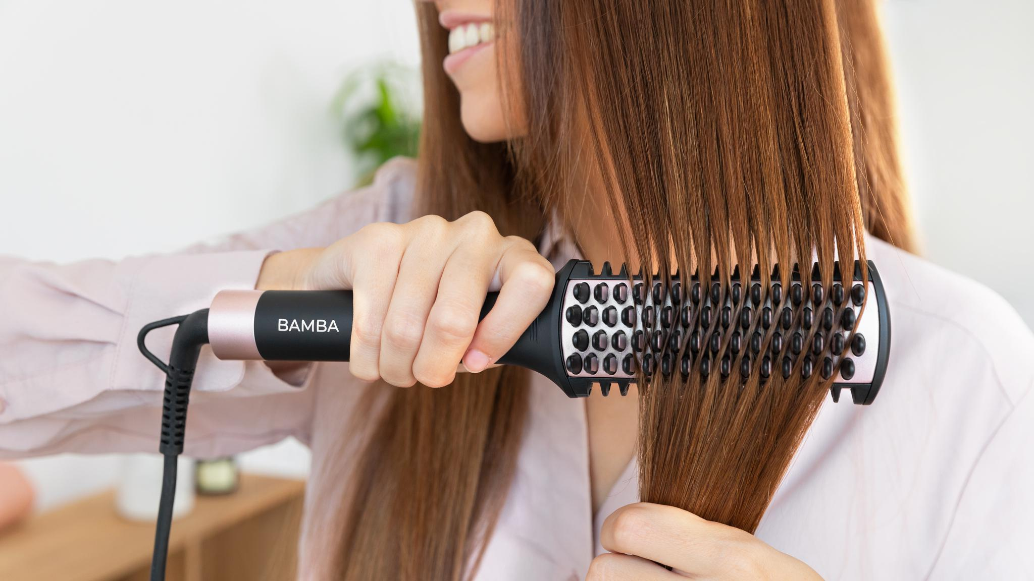 Una mujer se peina el cabello castaño y liso con un cepillo alisador eléctrico Bamba.