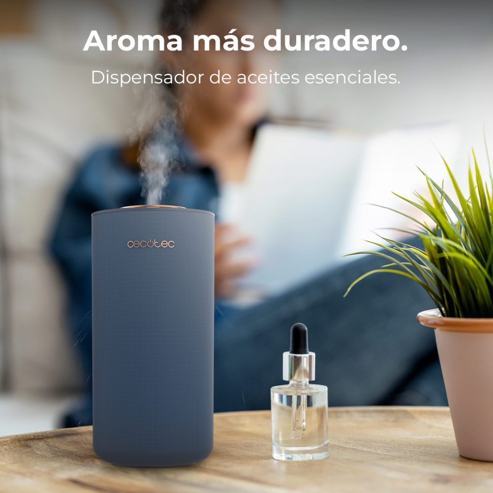 Un difusor azul expulsa vapor sobre una mesa de madera con un gotero y una planta; al fondo una mujer lee.