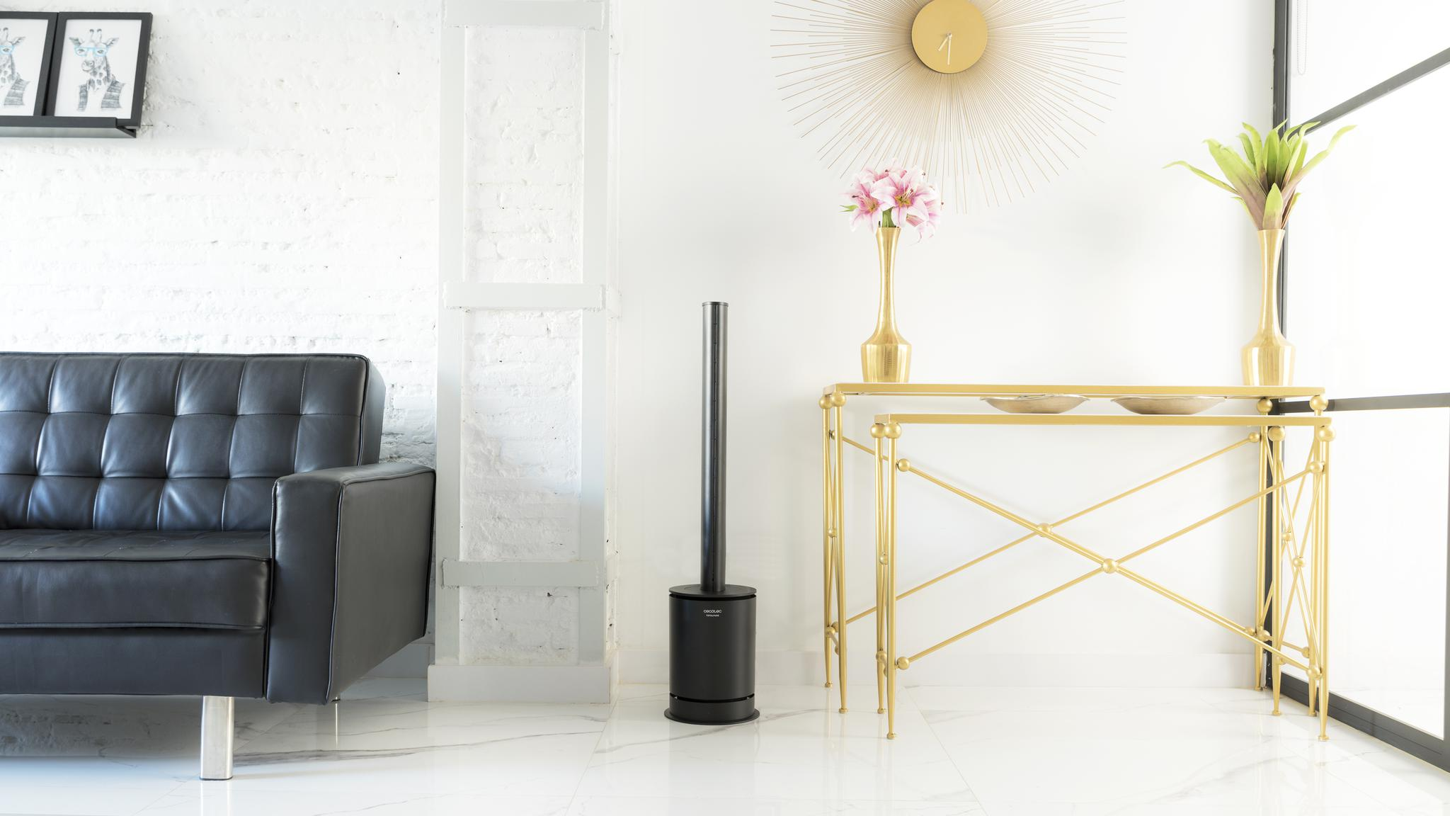 Un ventilador de torre negro se encuentra en una sala de estar luminosa junto a una mesa consola dorada y un sofá negro.