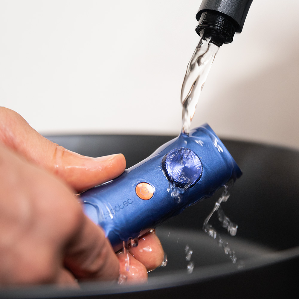 Una mano lava un dispositivo electrónico azul bajo agua corriente de una boquilla en un lavabo oscuro.