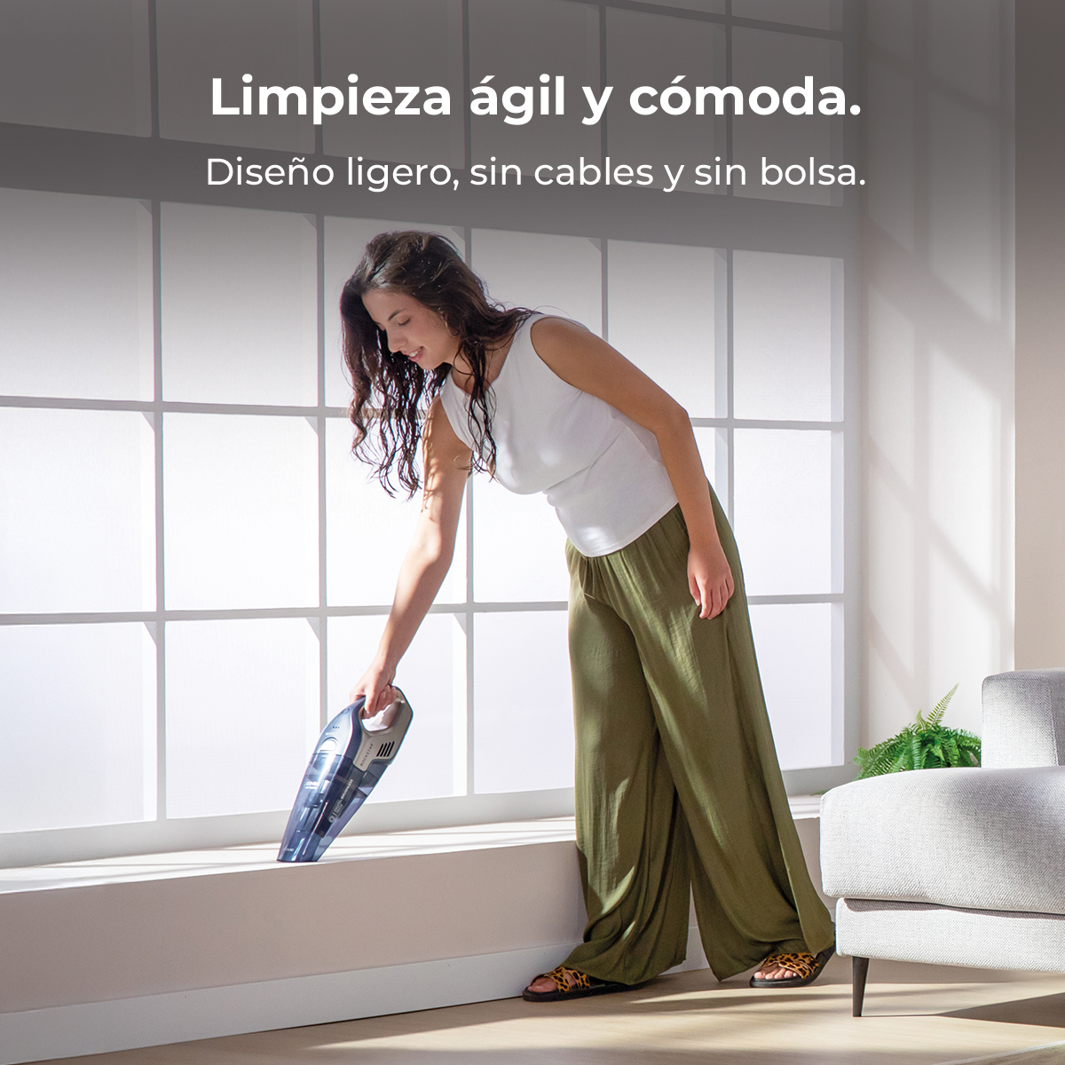 Una mujer sonriente con blusa blanca usa una aspiradora de mano en una repisa.