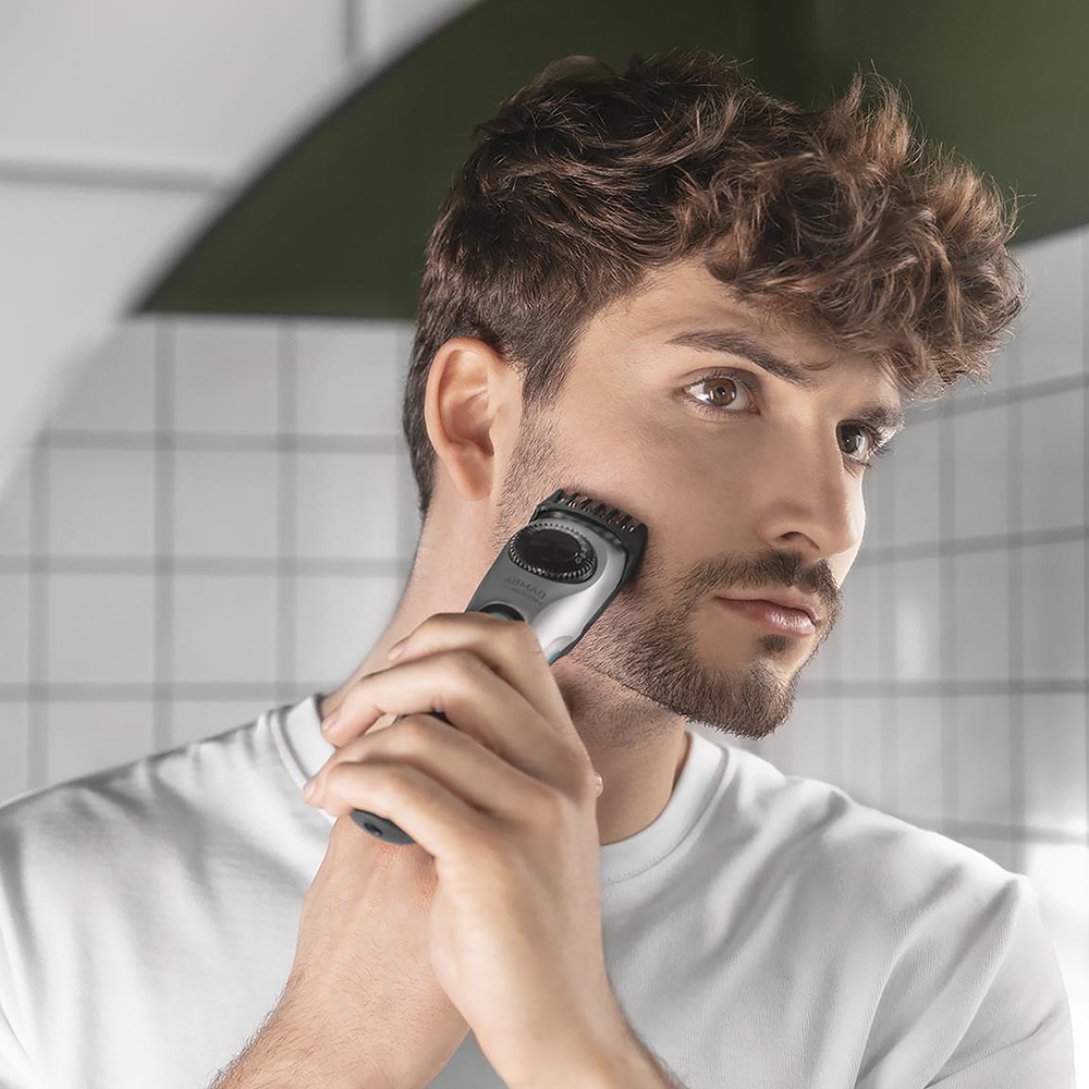 Un joven de cabello castaño se afeita la barba en un baño con azulejos blancos.