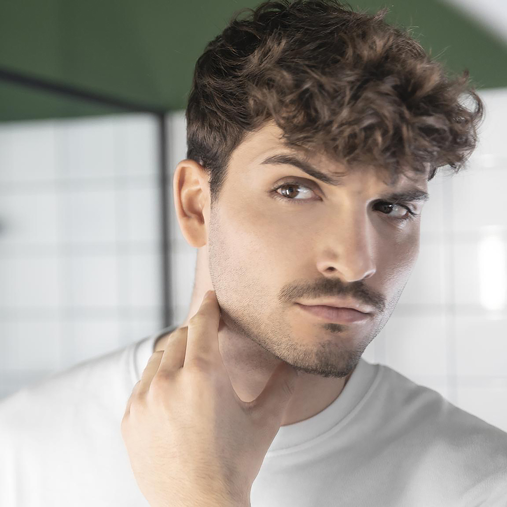 Un joven de pelo castaño corto y rizado y barba toca su cuello, con una camiseta blanca ante un muro alicatado.