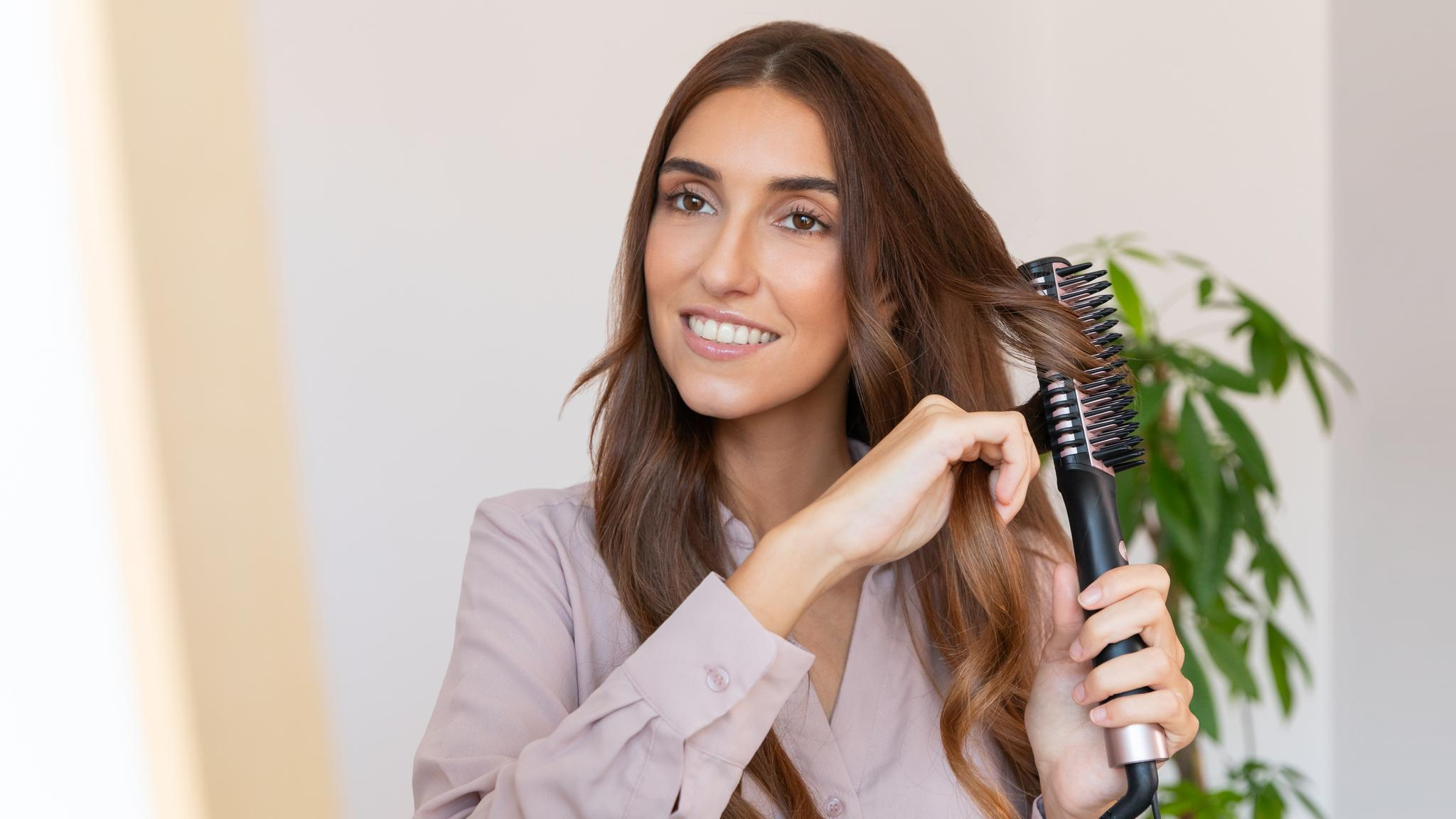 Una mujer sonriente se cepilla el pelo castaño largo con un cepillo alisador eléctrico en una habitación luminosa.