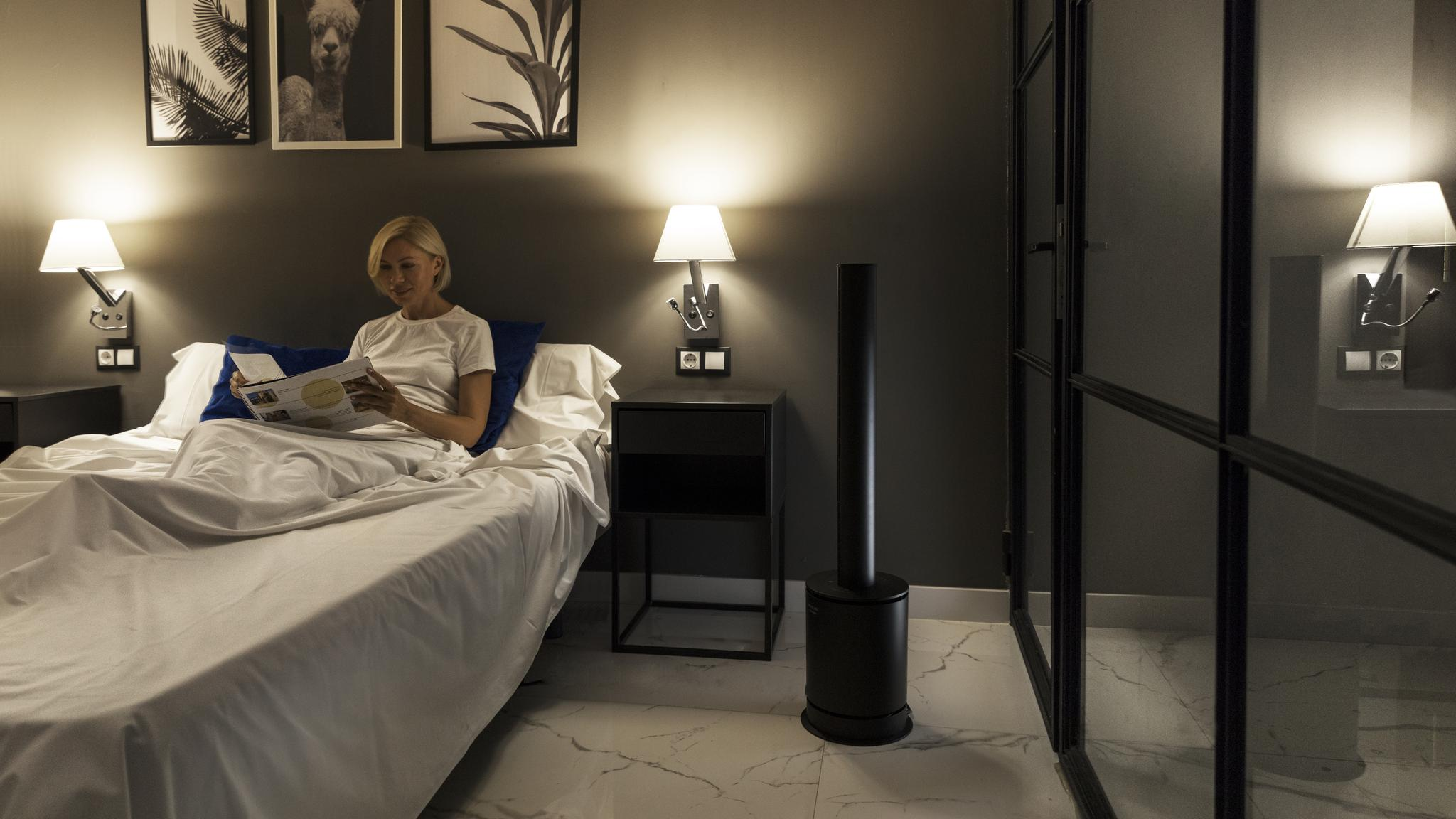 Una mujer rubia sentada en la cama lee una revista en un dormitorio poco iluminado con cuadros y un purificador.