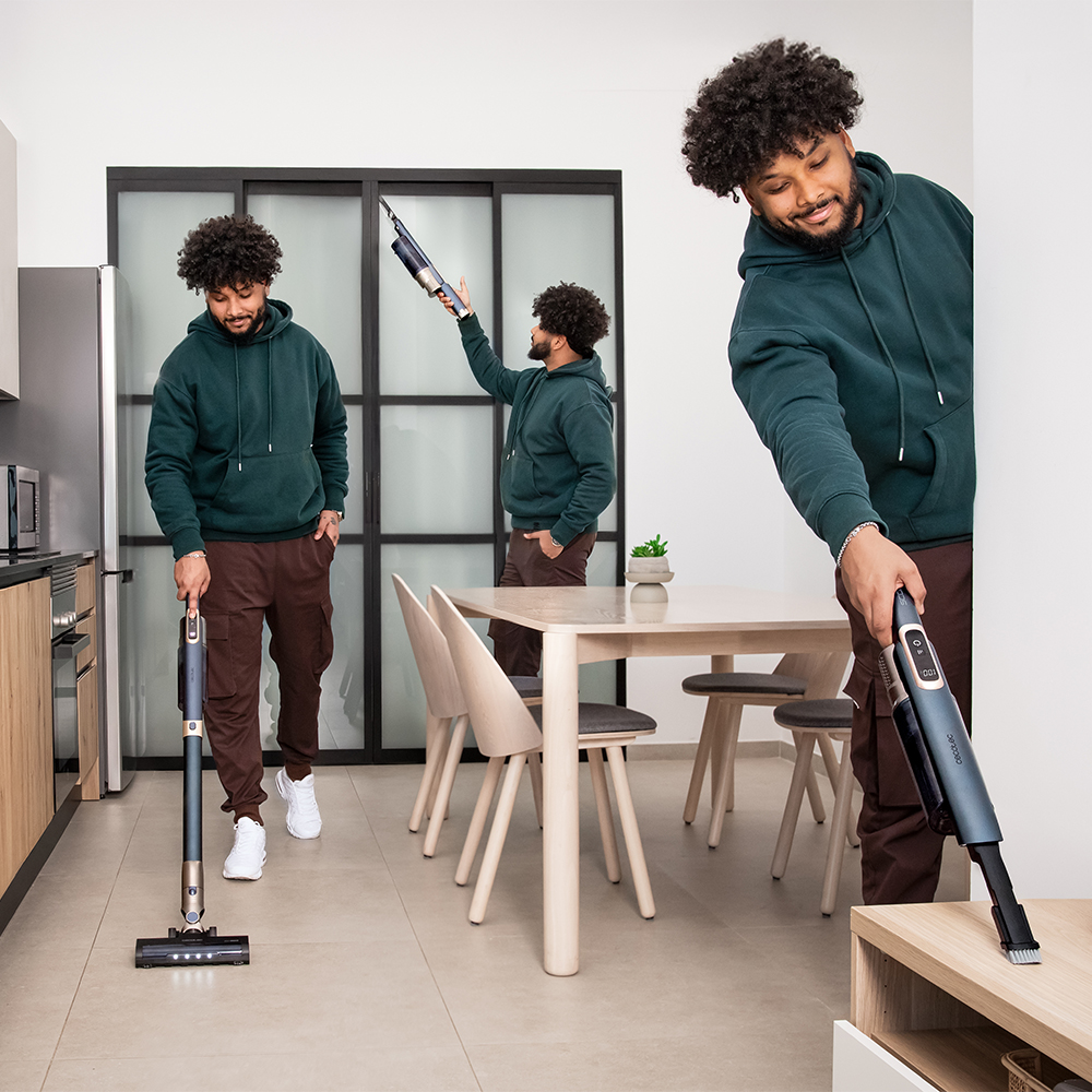 Un hombre con sudadera verde aspira varias zonas de un apartamento luminoso en una secuencia.