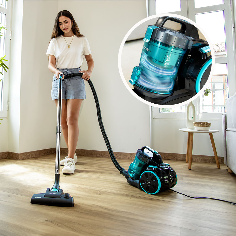 Una mujer aspira un suelo de madera con una aspiradora negra y turquesa, con un detalle circular mostrando su acción ciclónica.