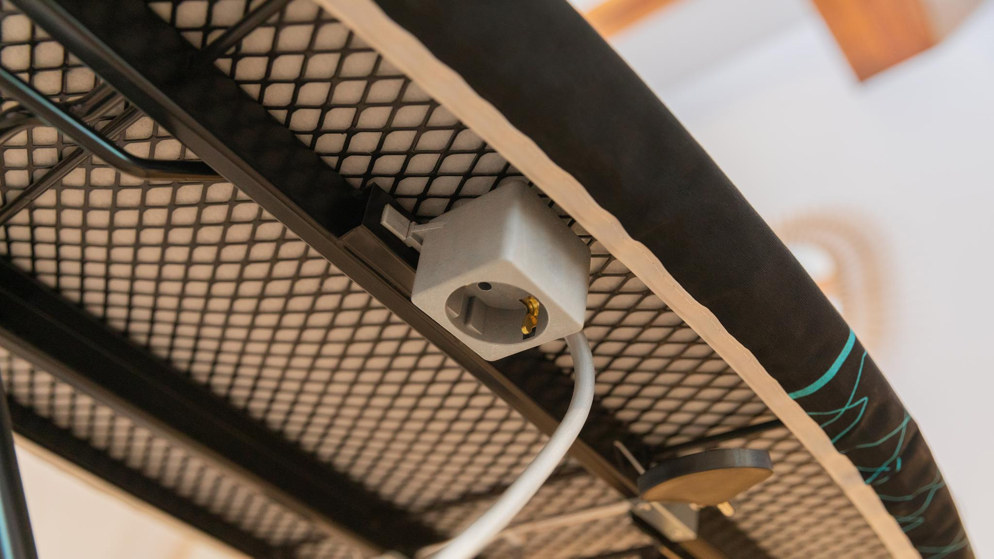 Parte inferior de una tabla de planchar con enchufe gris y cable blanco.