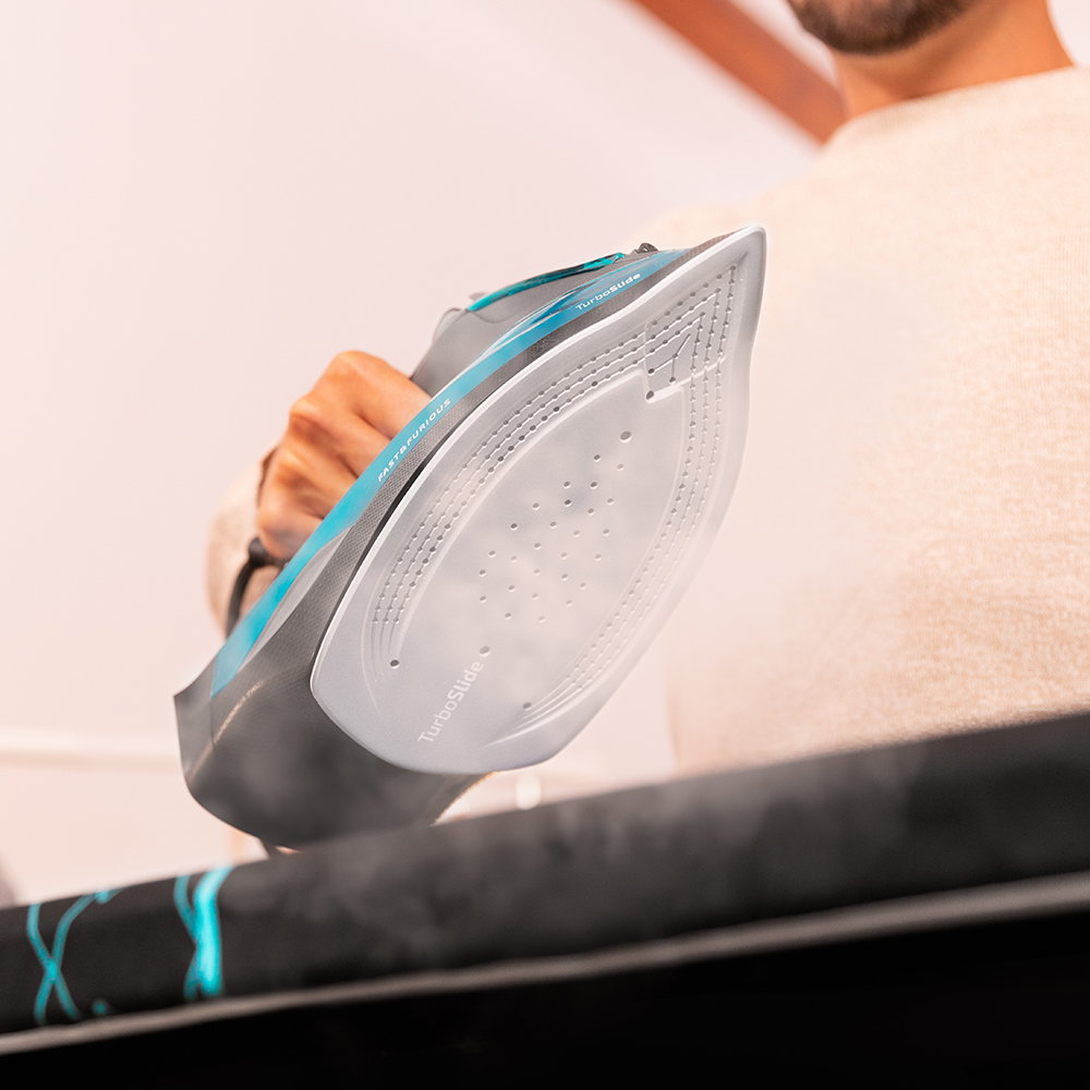 Un hombre sostiene una moderna plancha de vapor azul y gris sobre una tabla, mientras sale vapor.