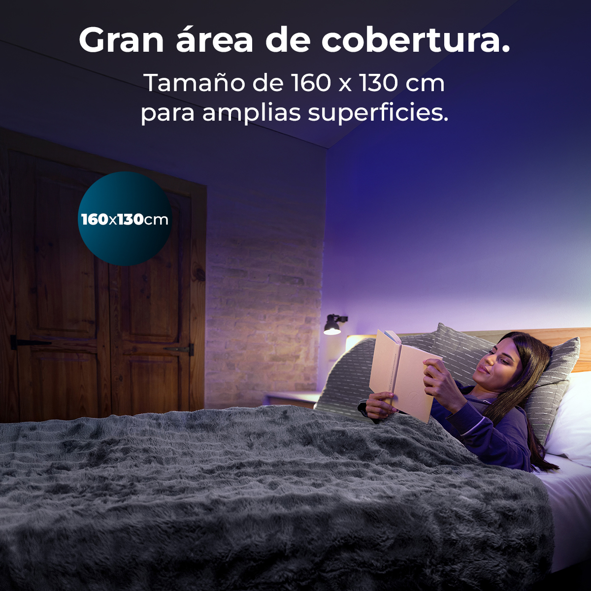 Una mujer lee un libro en la cama bajo una gran manta gris.