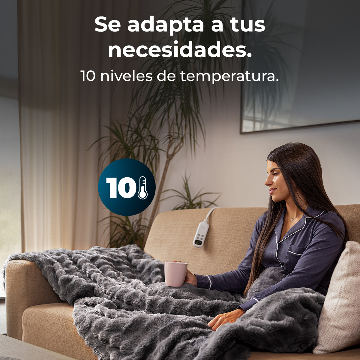 Una mujer se sienta en un sofá envuelta en una manta gris y suave, sosteniendo una taza.