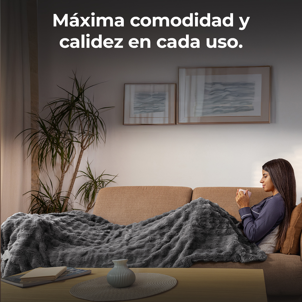 Una mujer se sienta cómodamente en un sofá, envuelta en una manta gris, sosteniendo una taza.