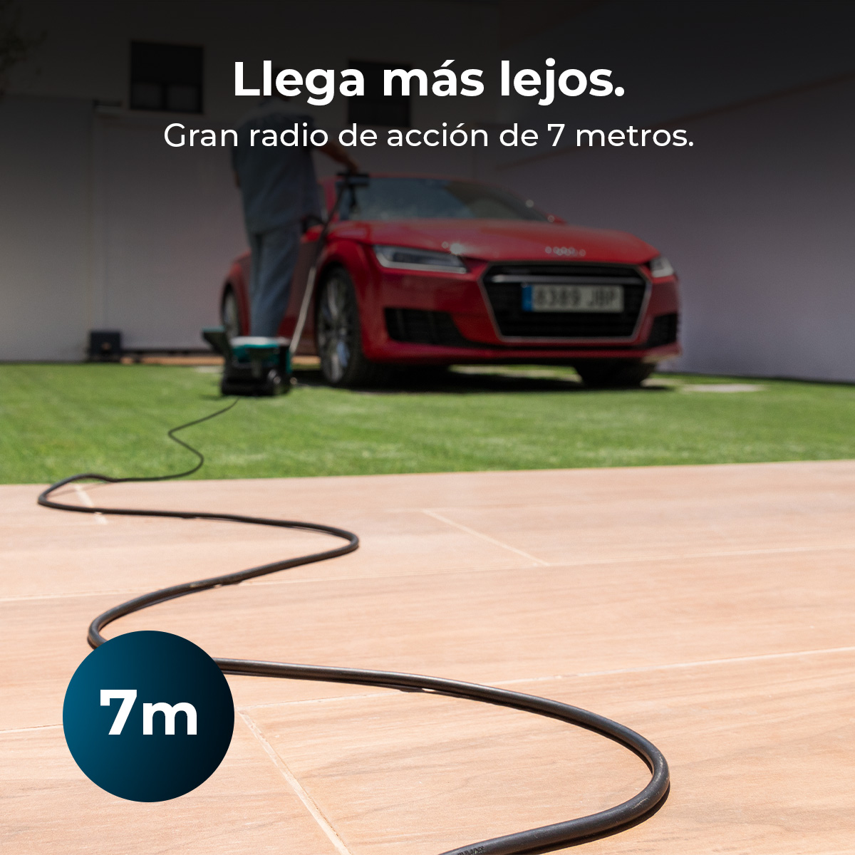 Una larga manguera negra de hidrolimpiadora cruza un patio de baldosas; al fondo, un coche rojo.