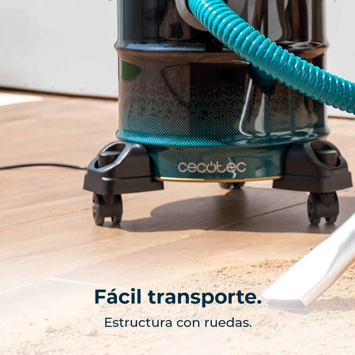 Aspiradora negra y turquesa con ruedas y manguera azul, aspirando suciedad en suelo de madera.