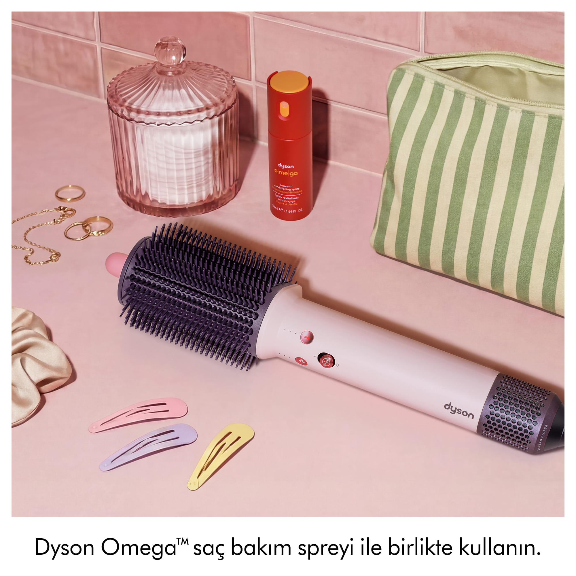 Pembe yüzey üzerinde aksesuarlar, saç tokaları, sprey ve çizgili çanta ile bir Dyson saç şekillendirici.