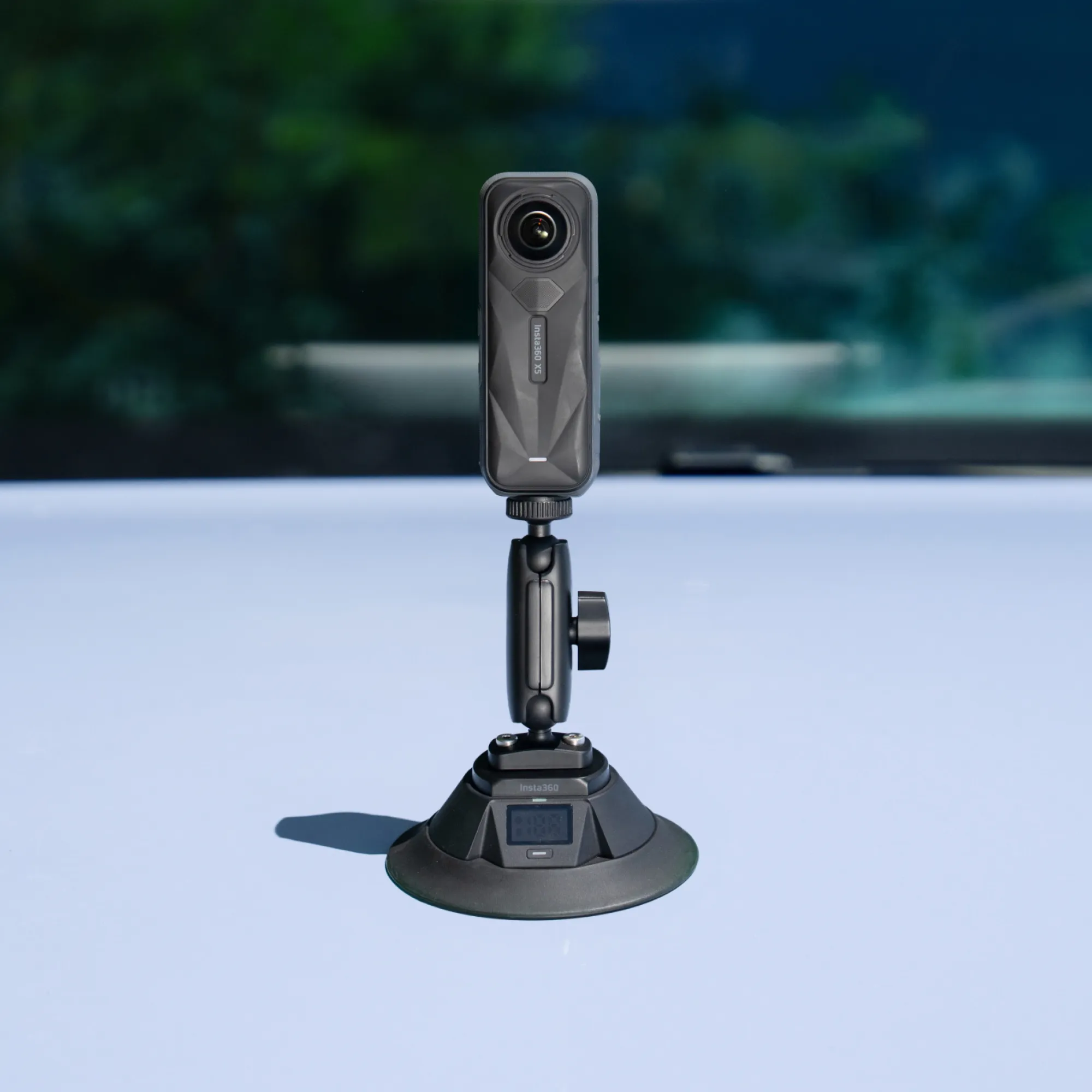 Fekete Insta360 akciókamera tapadókorongos állványon egy autó motorháztetőjén, elmosódott zöld háttérrel.