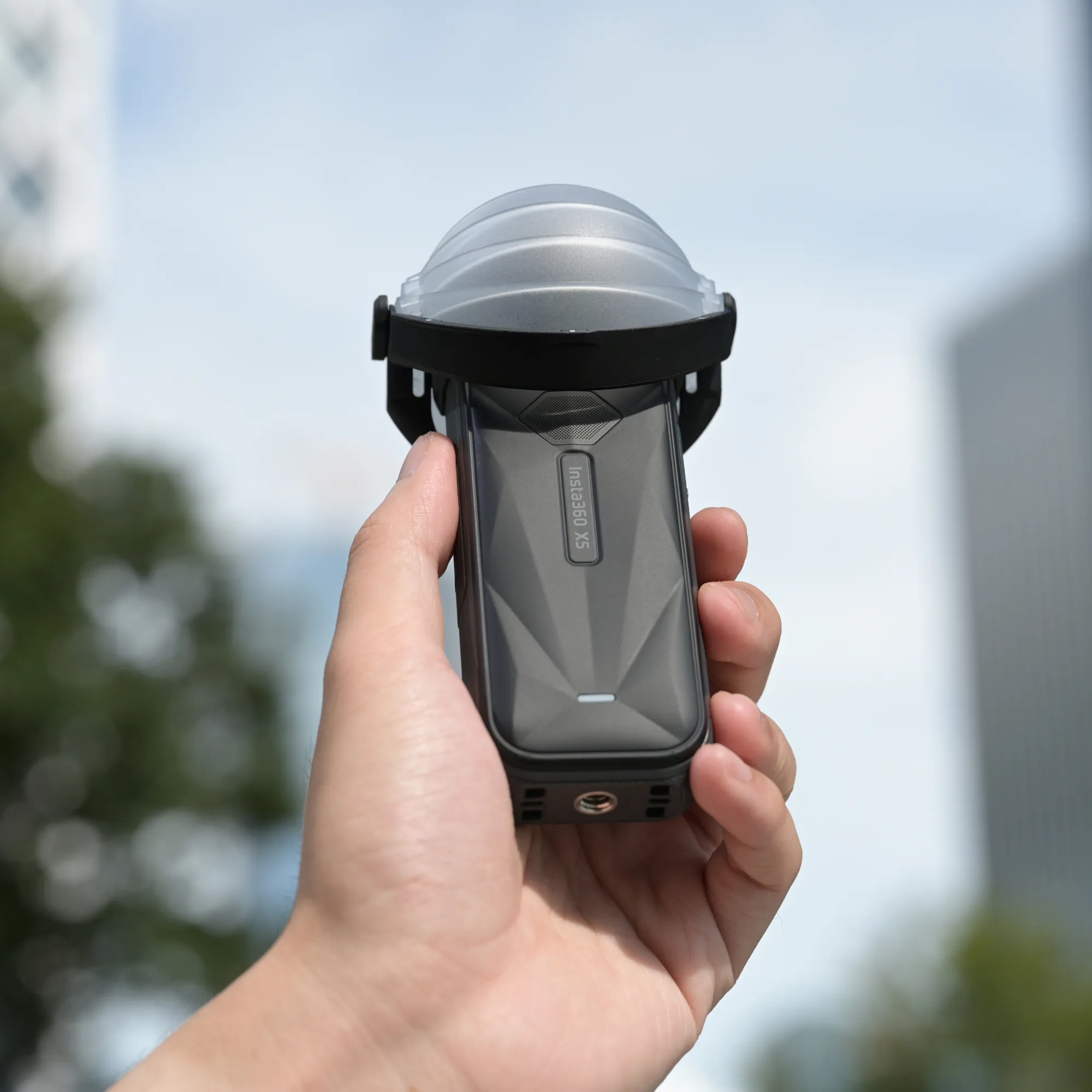 Egy kéz egy sötét Insta360 X5 kamerát tart védőtokban, elmosódott háttér előtt.