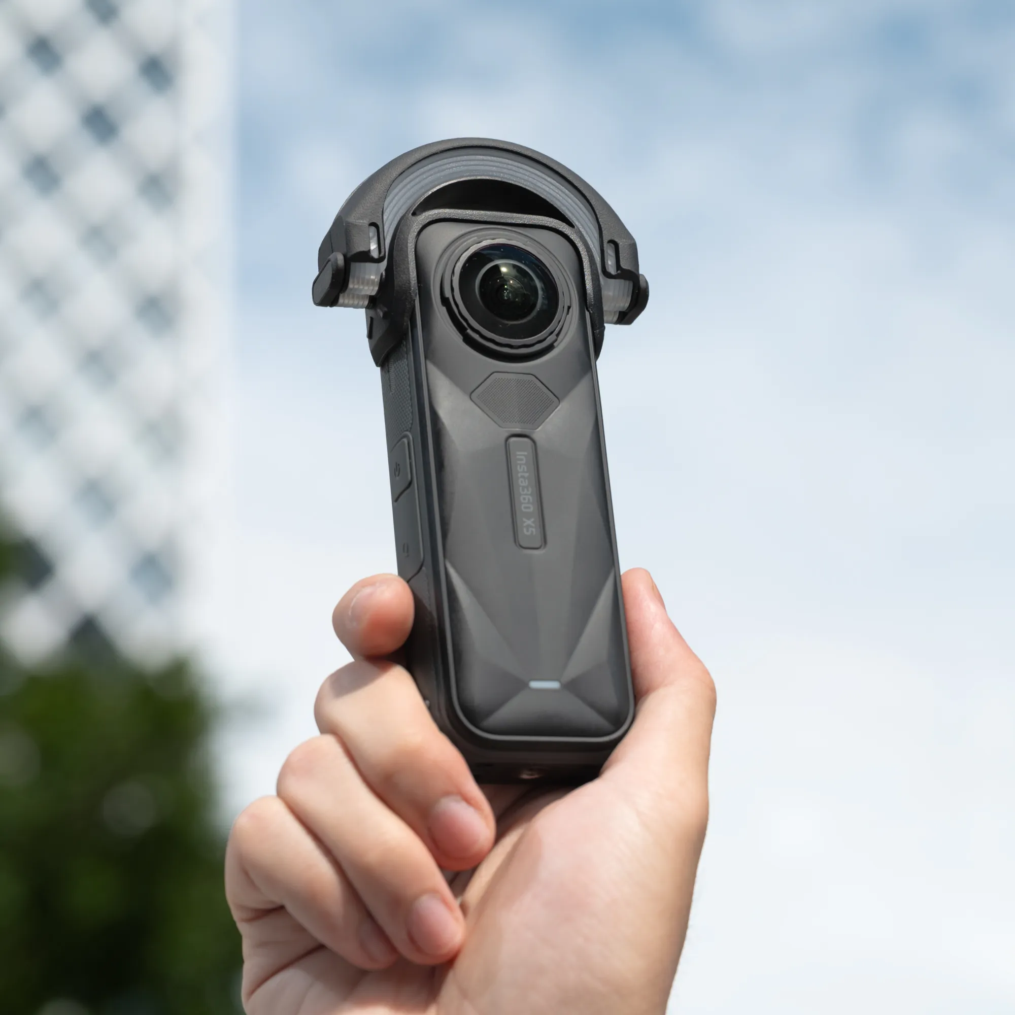 Egy kéz egy fekete Insta360 X3 kamerát tart az égbolt és épületek elmosódott háttere előtt.
