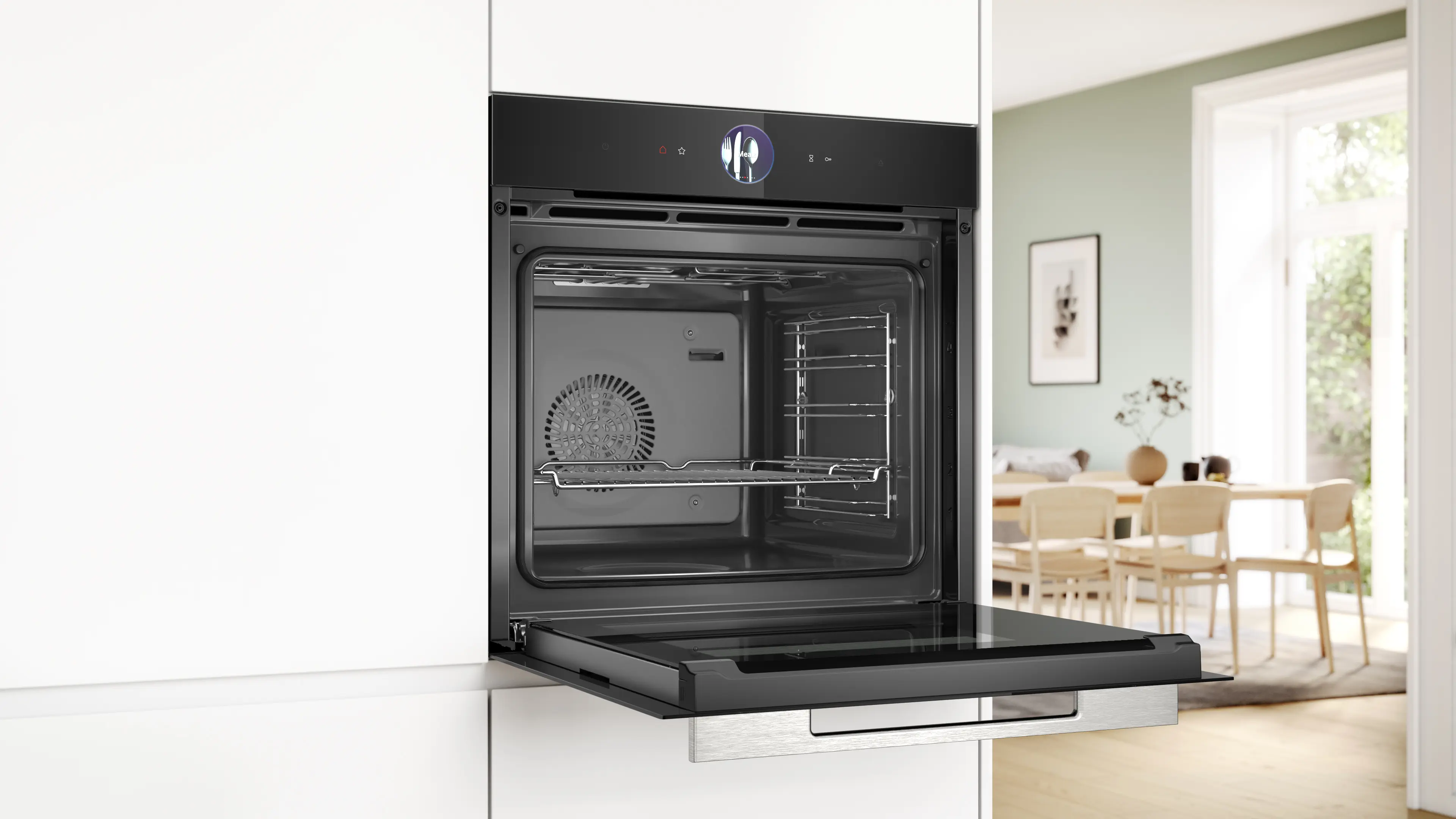 Horno negro con puerta abierta en cocina blanca, con un comedor iluminado visible al fondo.