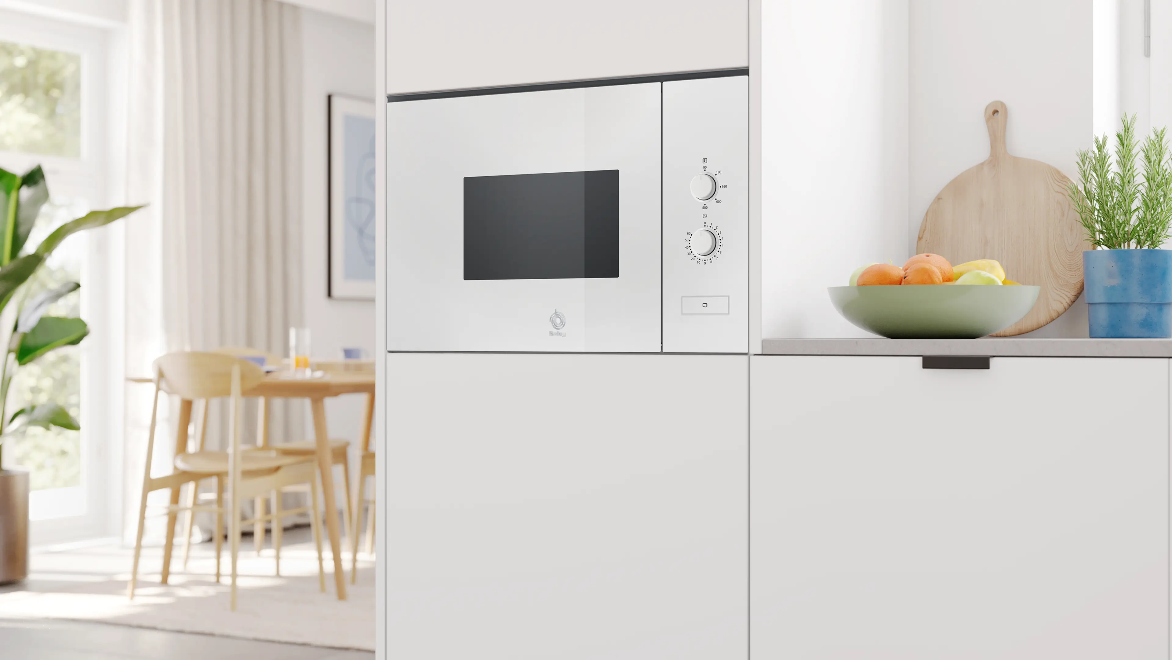 Un microondas blanco está empotrado en los muebles de cocina, con un frutero y hierbas al lado.
