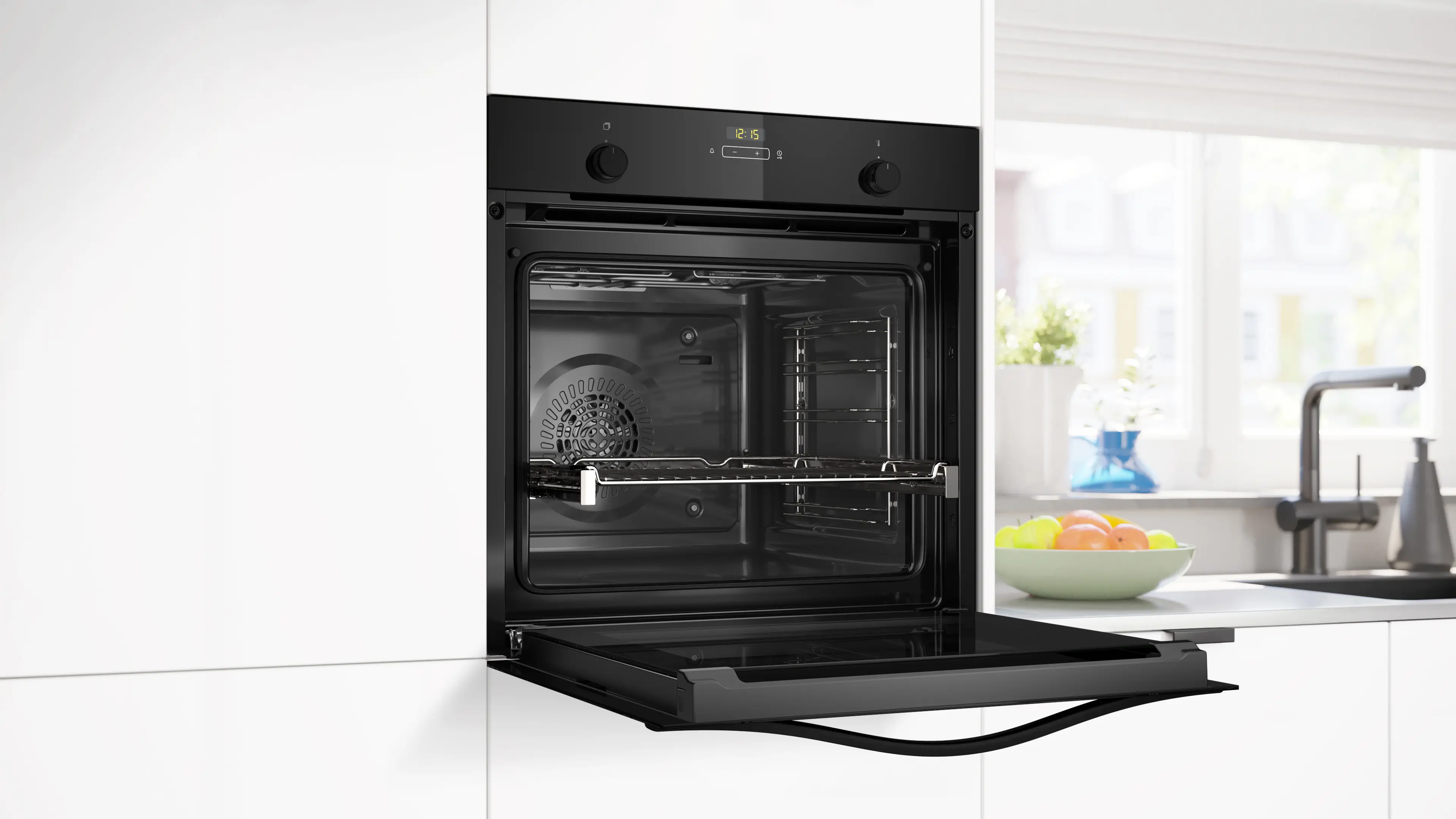 Un horno empotrado negro con puerta abierta en cocina blanca, con frutero y ventana al fondo.