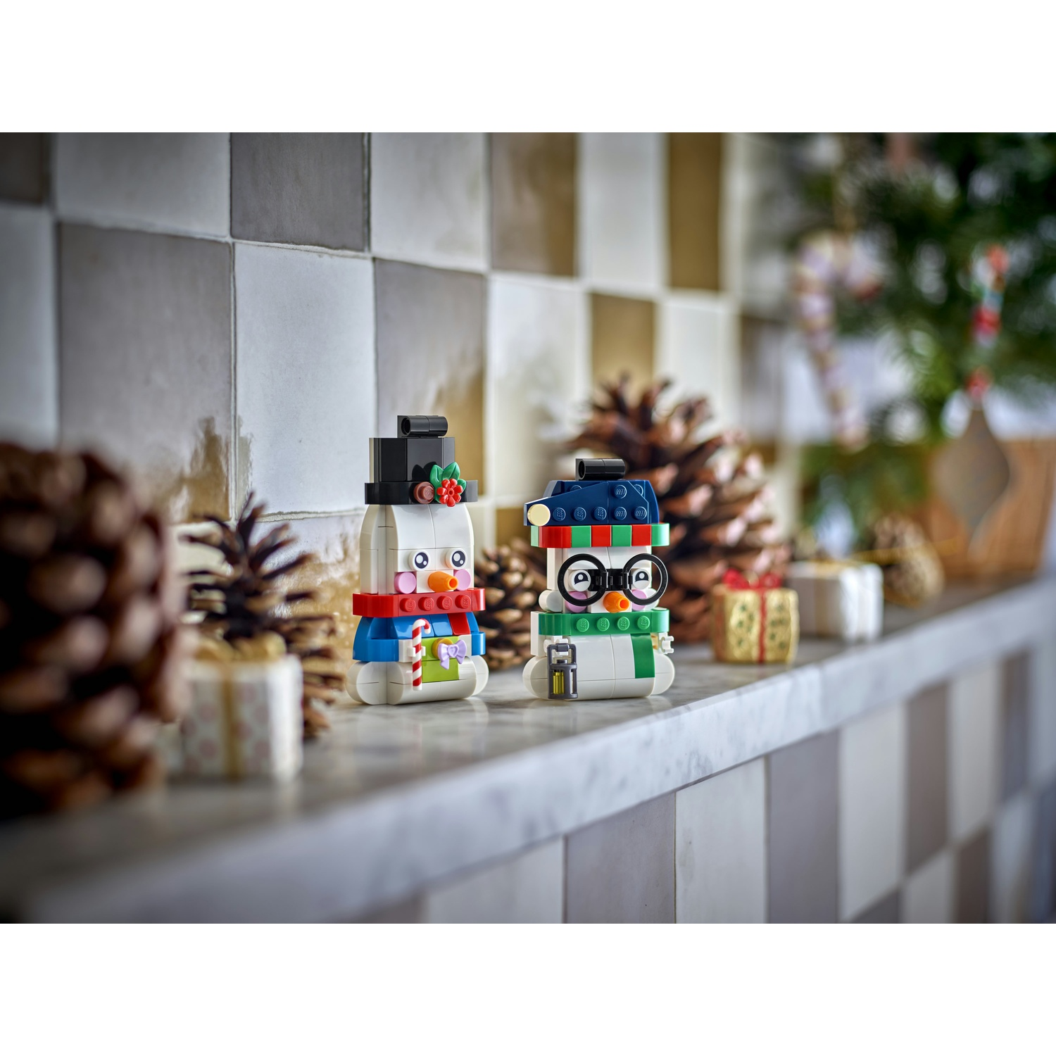 Due piccoli pupazzi di neve Lego sono su una mensola, circondati da pigne e regali.