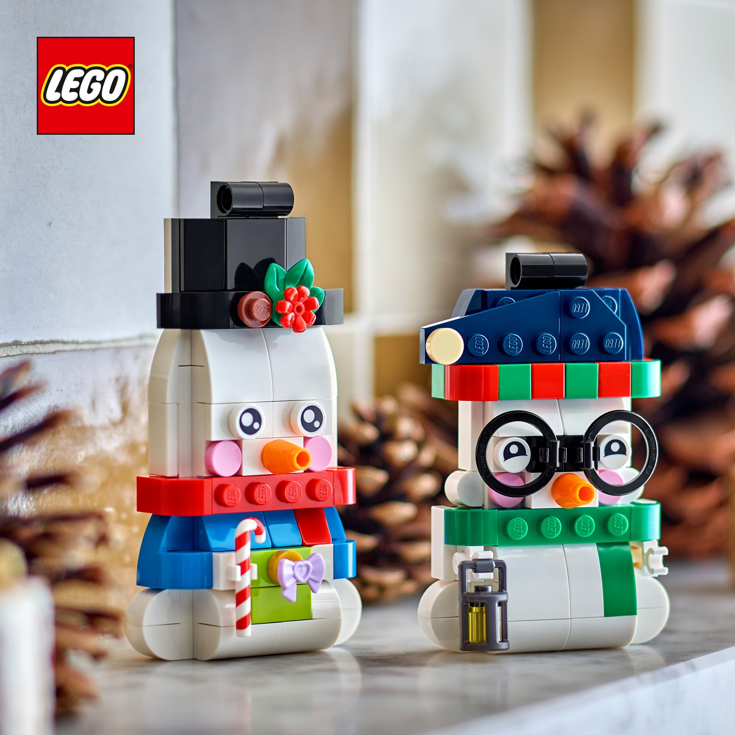 Due statuette di pupazzi di neve Lego si trovano su una superficie, circondate da pigne, con il logo Lego.